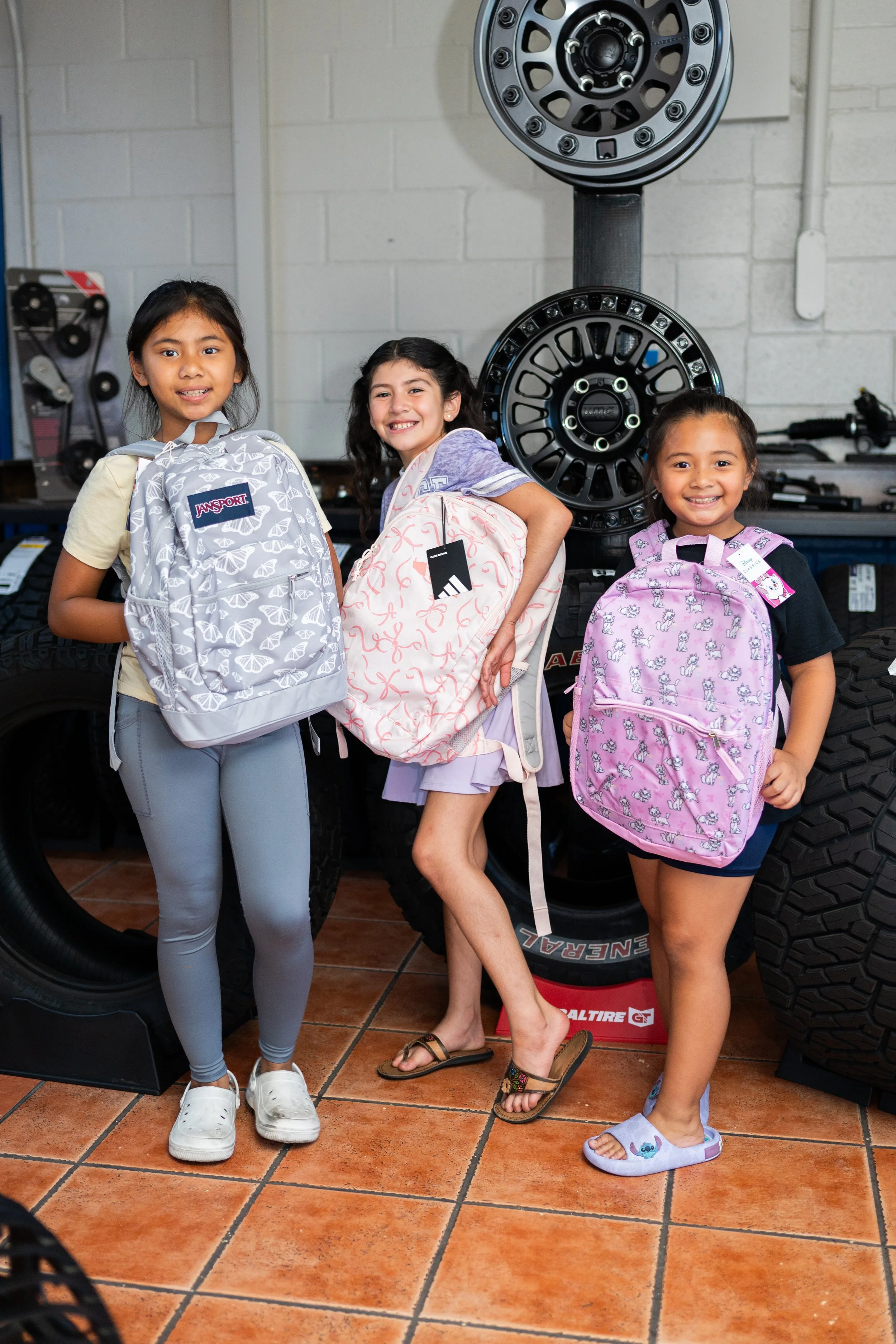 Three young girls standing inside an auto parts store, each holding a backpack and smiling at the camera. Behind them are car tires and rim displays.
