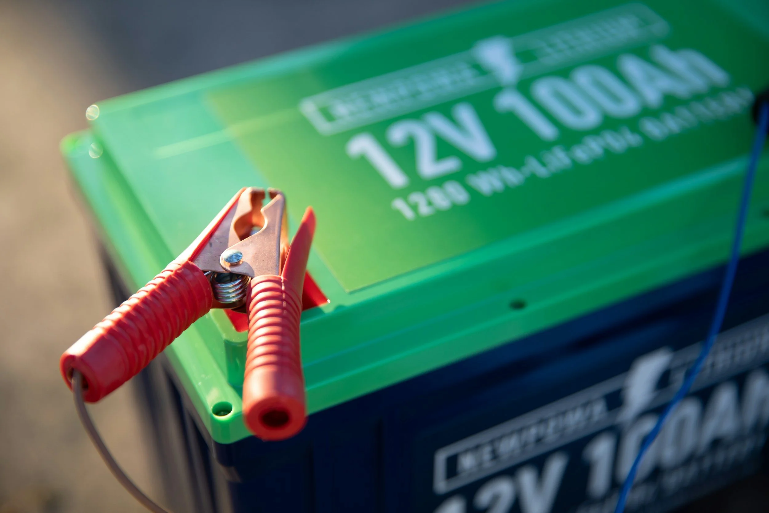 Close-up of a hand holding a clipper connected to orange and gray wires, with a blurred green electric vehicle charging station in the background.