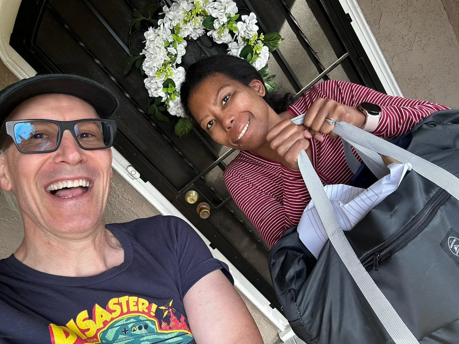 A man and a woman taking a selfie in front of a door with a white floral wreath. The man is smiling and wearing glasses and a black cap, and the woman is smiling, holding a strap, and has a backpack beside her.