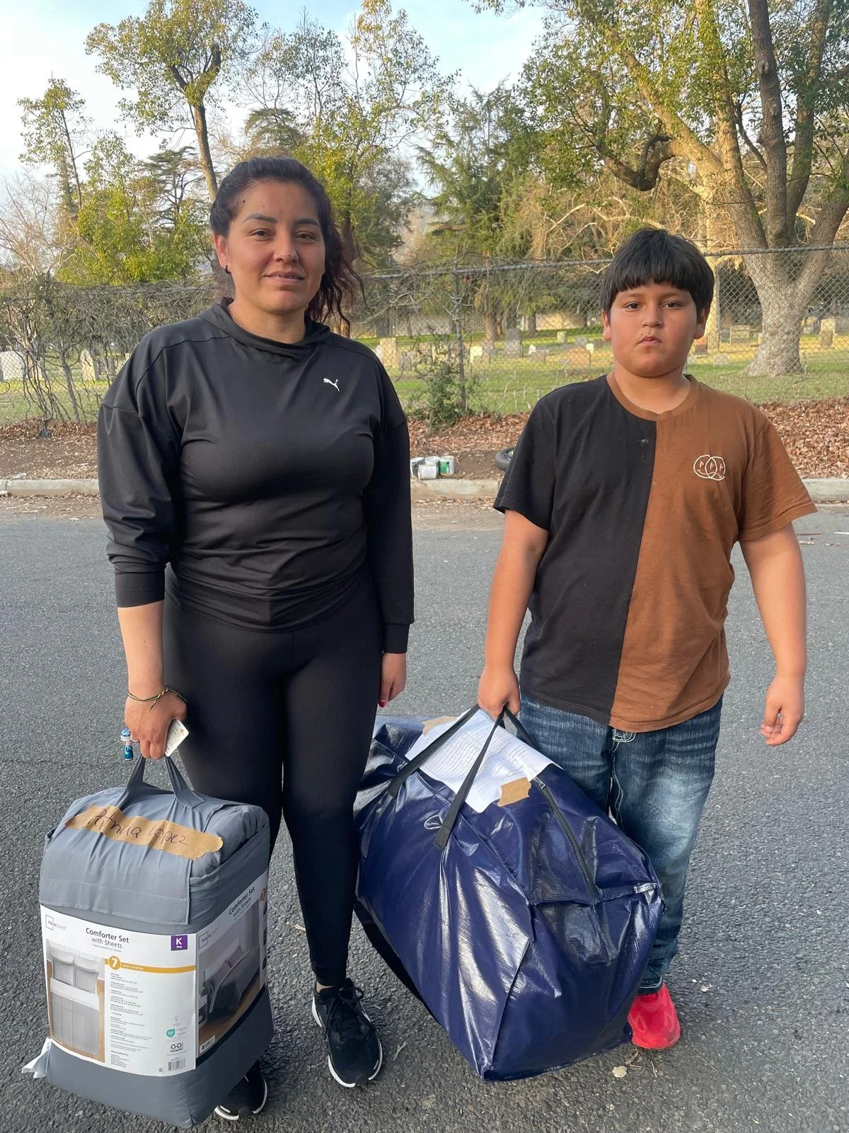A woman and a young boy standing outdoors on a paved area, holding large bags filled with items, with trees and a fence in the background.