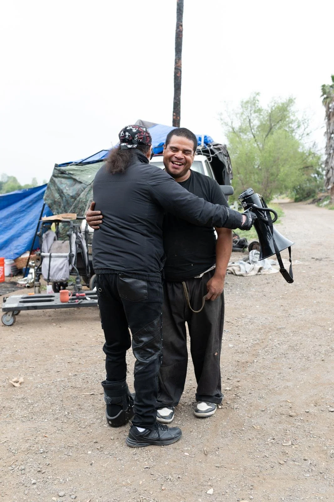 Two men hugging outdoors next to a makeshift shelter, one smiling with a megaphone in hand.