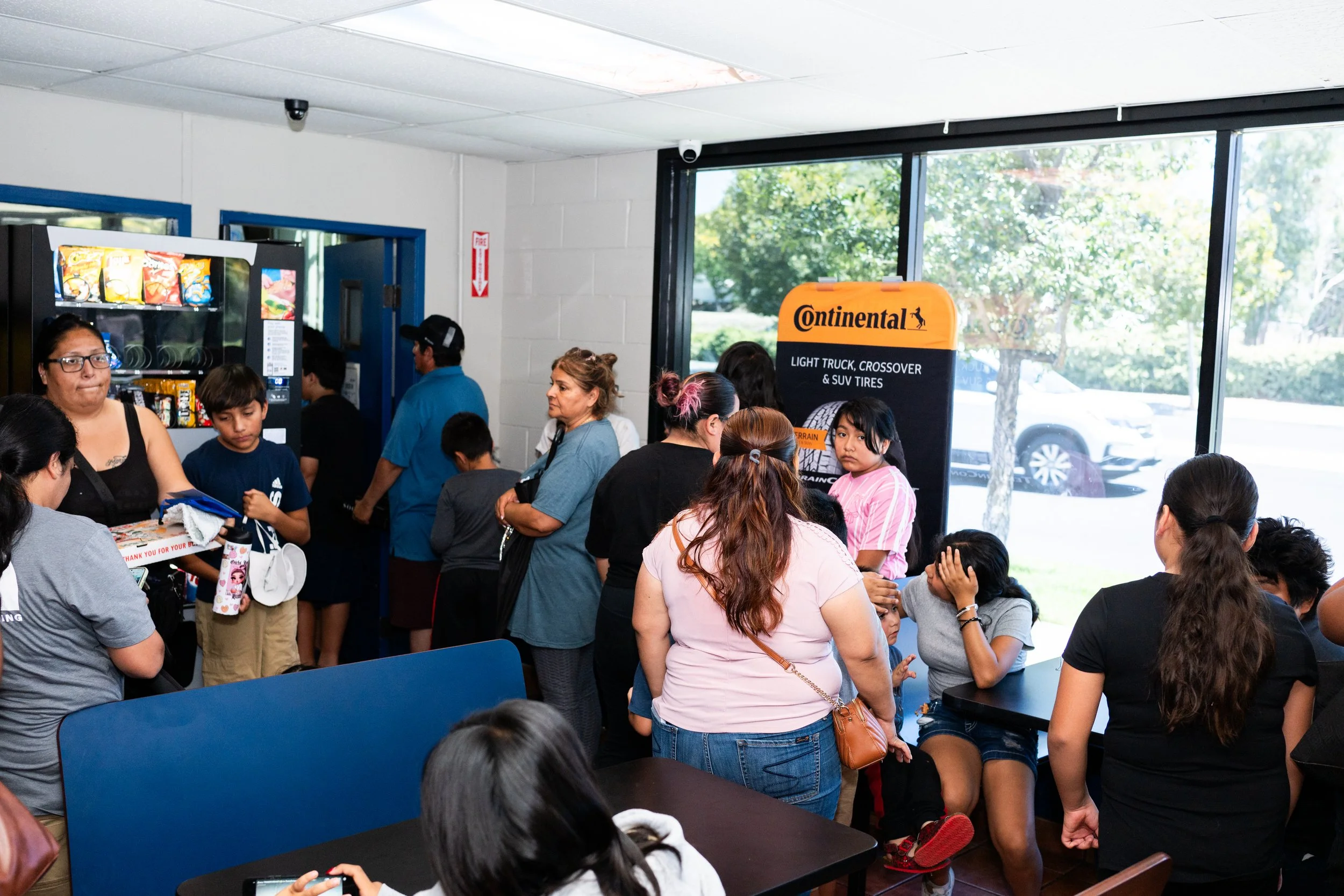 A crowded indoor waiting area with people standing and sitting, including children and adults. There is a vending machine with snacks and drinks, and a sign for Continental tires near a large window showing trees outside.