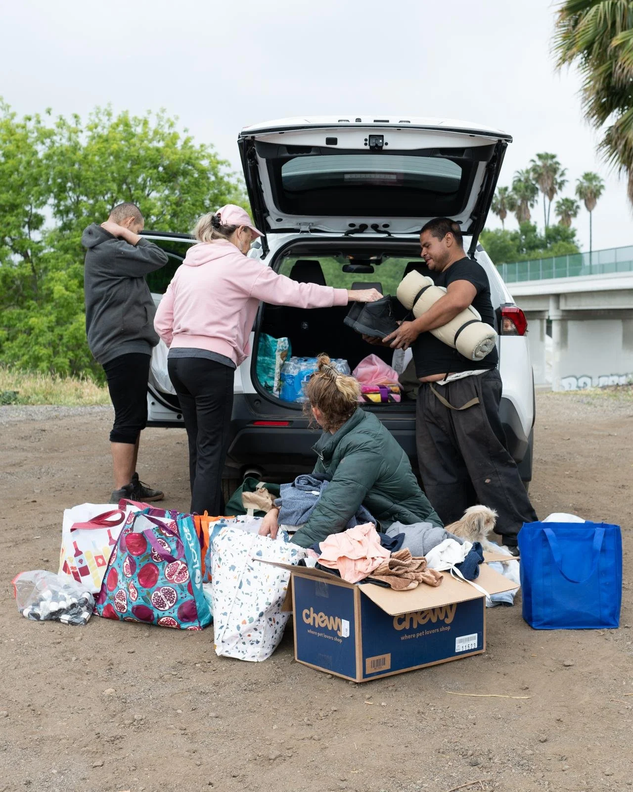 Family loading supplies into the back of a white SUV outdoors, with bags, boxes, and camping gear on the ground.
