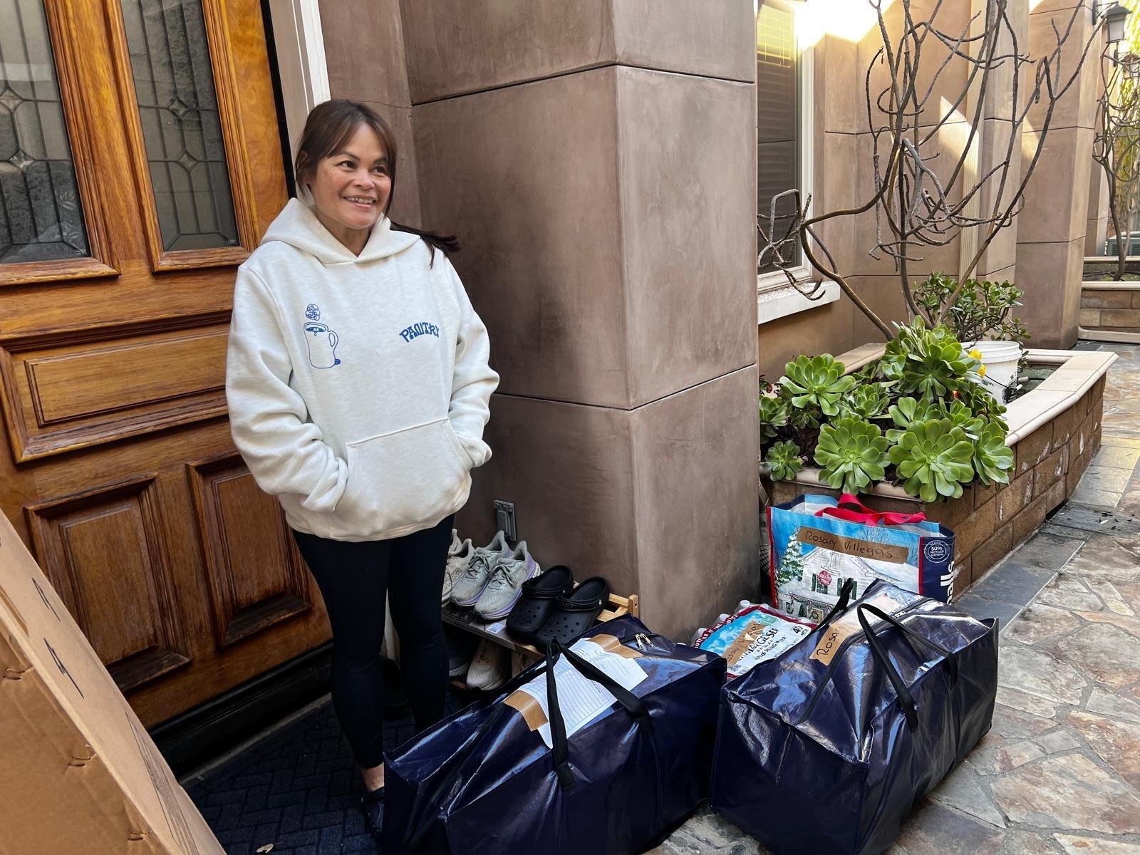 A woman standing outside a house with a smile, wearing a white hoodie, in front of a door, with various packages, shoes, and bags on the ground.