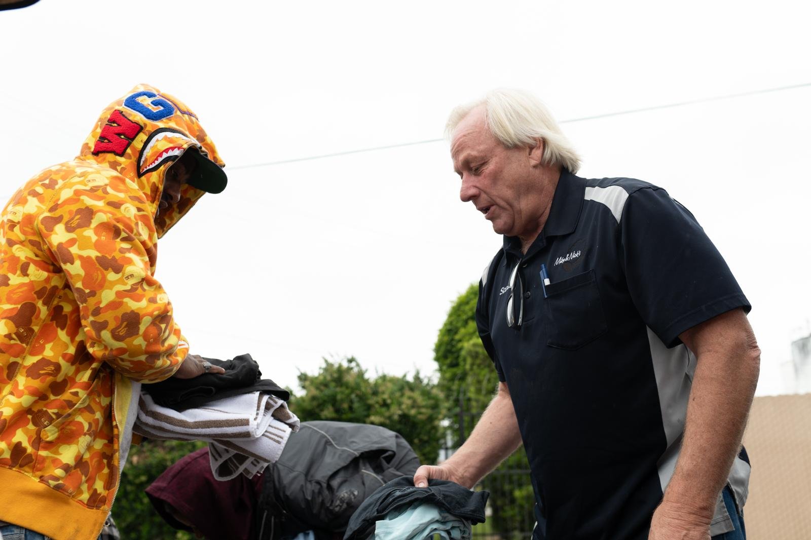 Two men are folding clothes outdoors on a cloudy day. One man is wearing a colorful camouflage hoodie with a shark face and the word 'WGM' on the hood. The other man has light hair and is dressed in a dark shirt with a name tag.