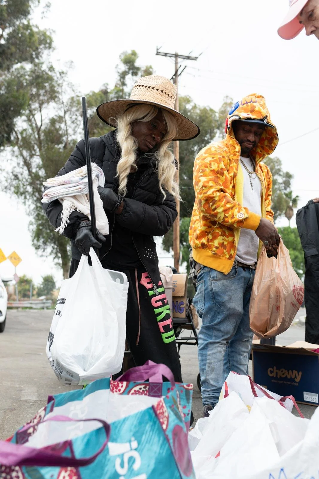 People at an outdoor event with bags and boxes, some holding plastic grocery bags, under a cloudy sky.