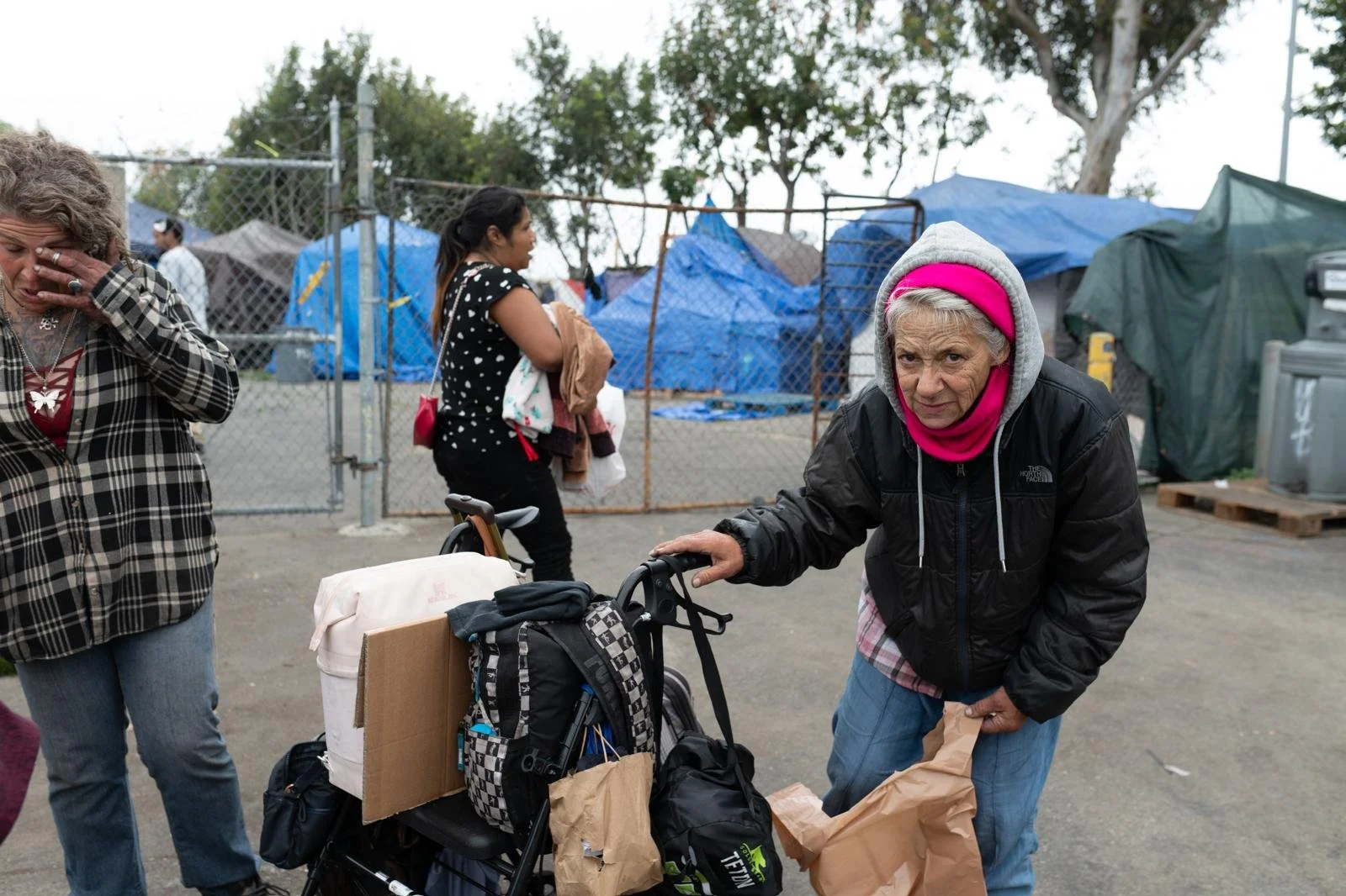 An elderly woman with a pink headband and gray hoodie pushes a shopping cart filled with bags and supplies outside a makeshift shelter area with blue tents and a chain-link fence.