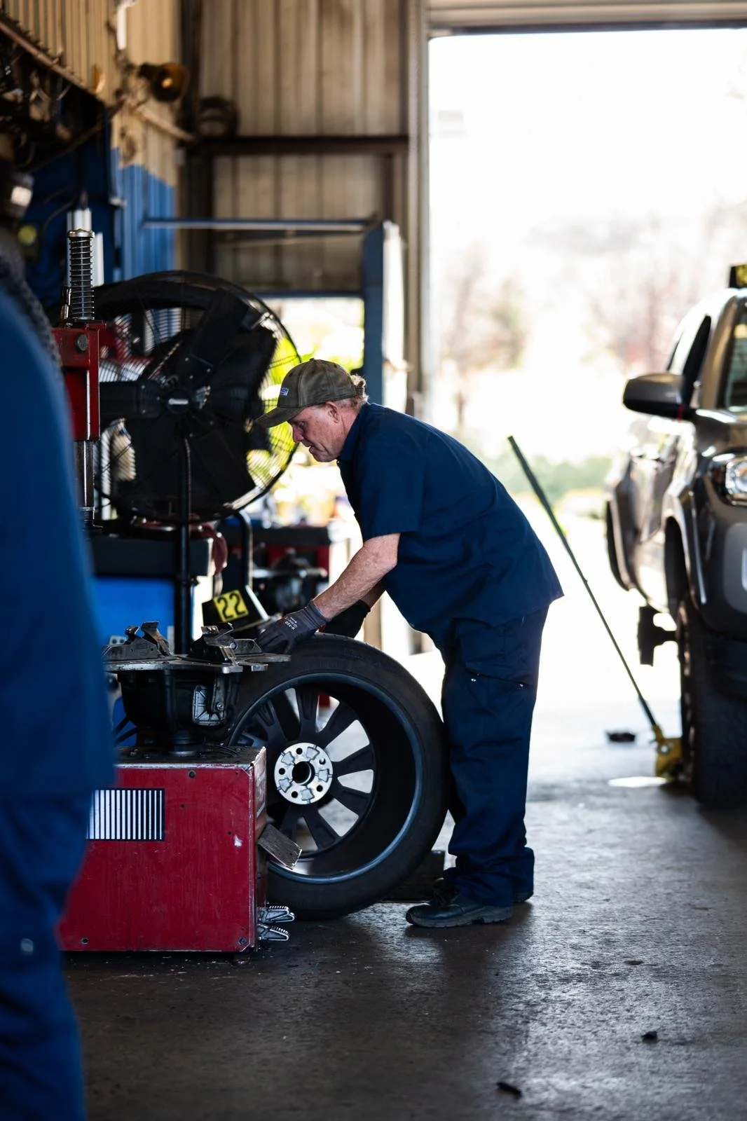 A mechanic working on a tire inside an auto repair shop, with cars and tools visible in the background.