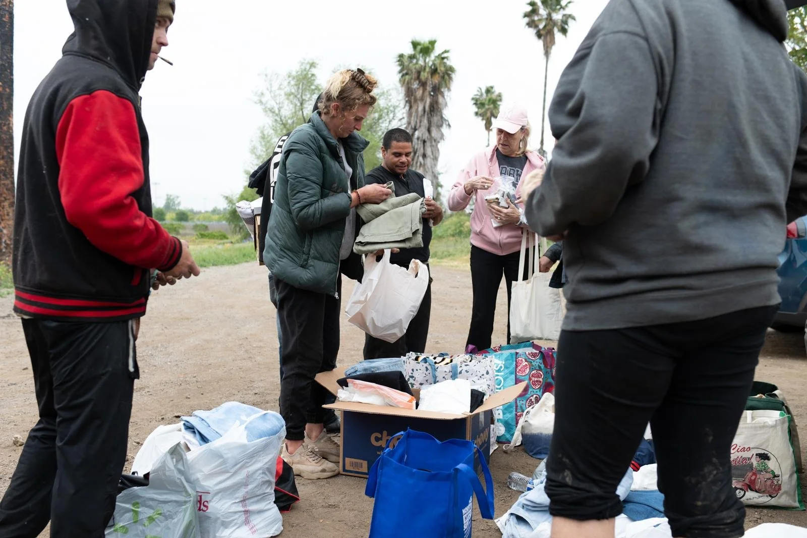 Group of people exchanging clothes and supplies outdoors during the daytime, with trees and palm trees in the background.
