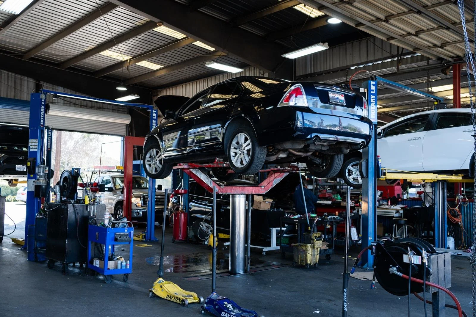 Inside an auto repair shop with a black sedan lifted on a two-post lift, various tools and equipment around, and a white car on another lift in the background.