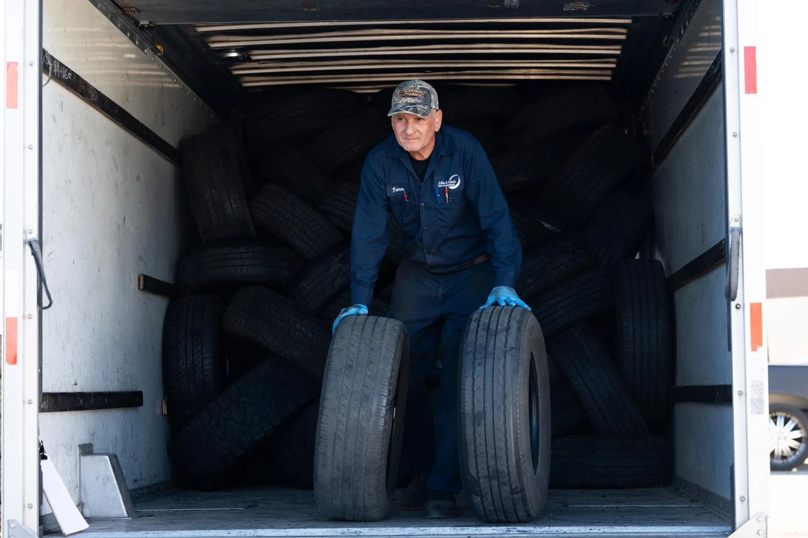 A man in a blue work uniform and cap unloading tires from a truck with a partially filled cargo area and many stacked tires inside.