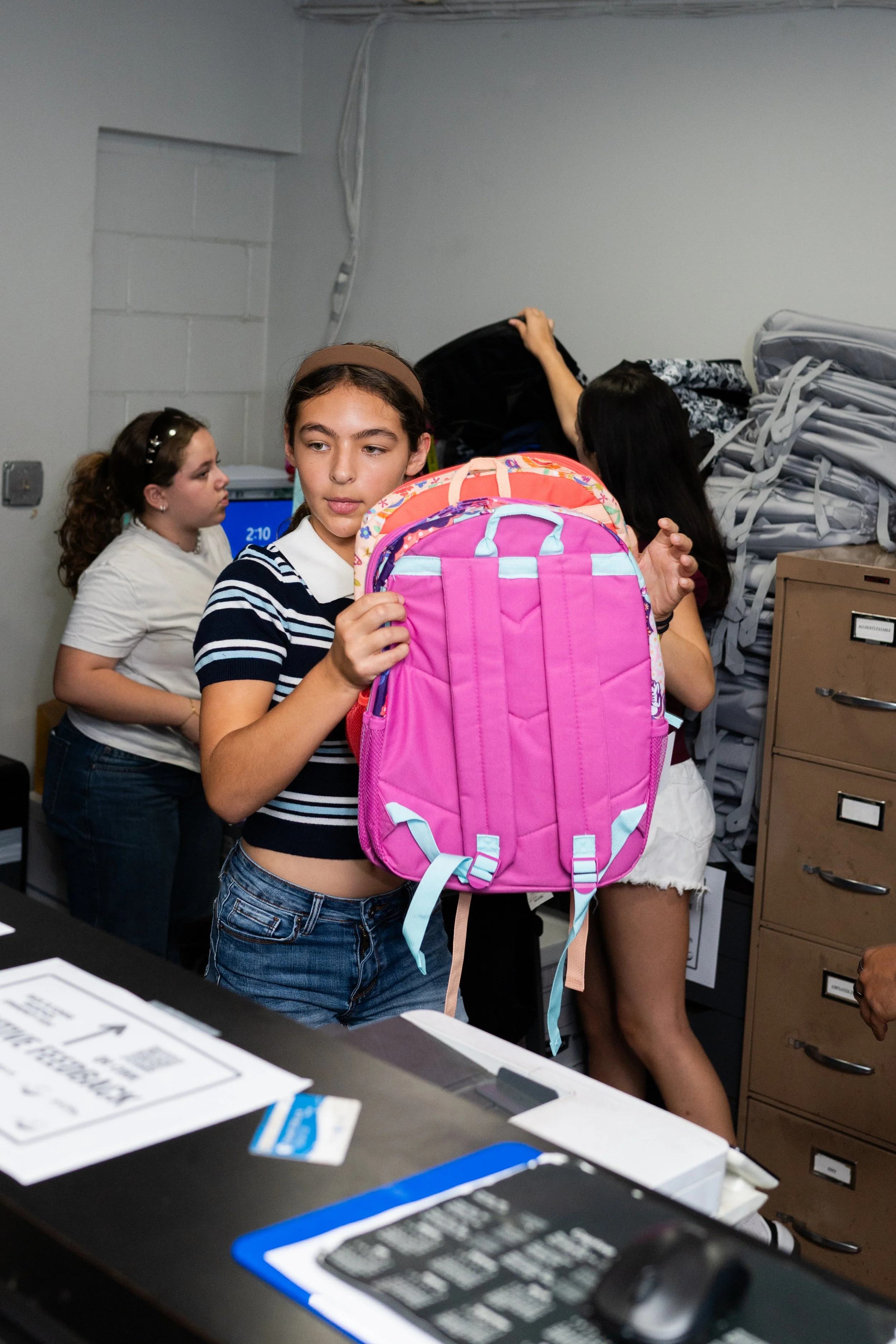 A girl in a striped shirt holding a pink backpack in a busy office or school setting with other girls in the background, some handling bags and a computer monitor showing 2:10.