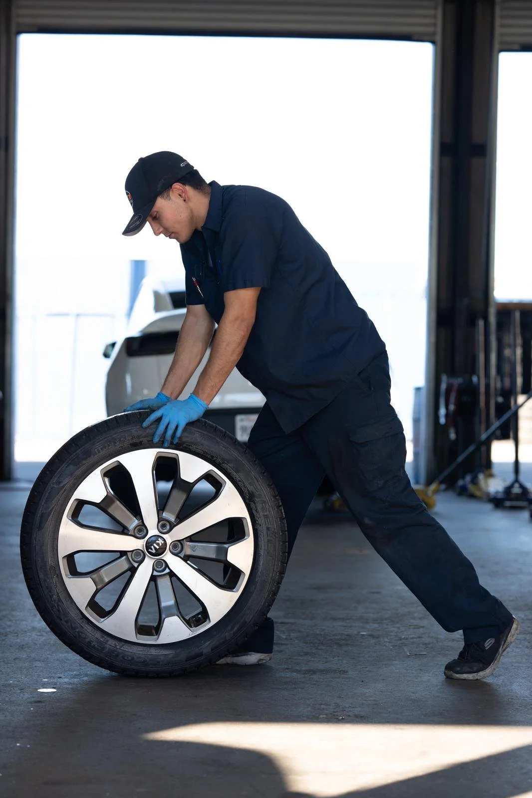 A mechanic wearing a black uniform, gloves, and a cap is flipping a car tire inside a garage with a car in the background.