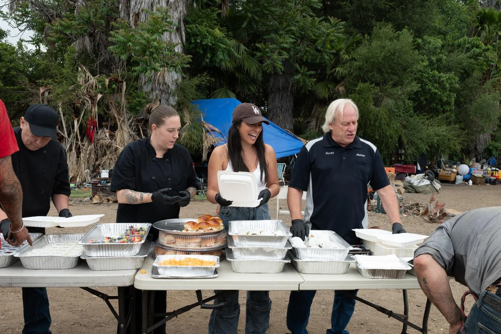 People preparing food at an outdoor gathering, with tables filled with trays of food, buns, and toppings, in a natural setting with trees and tents in the background.