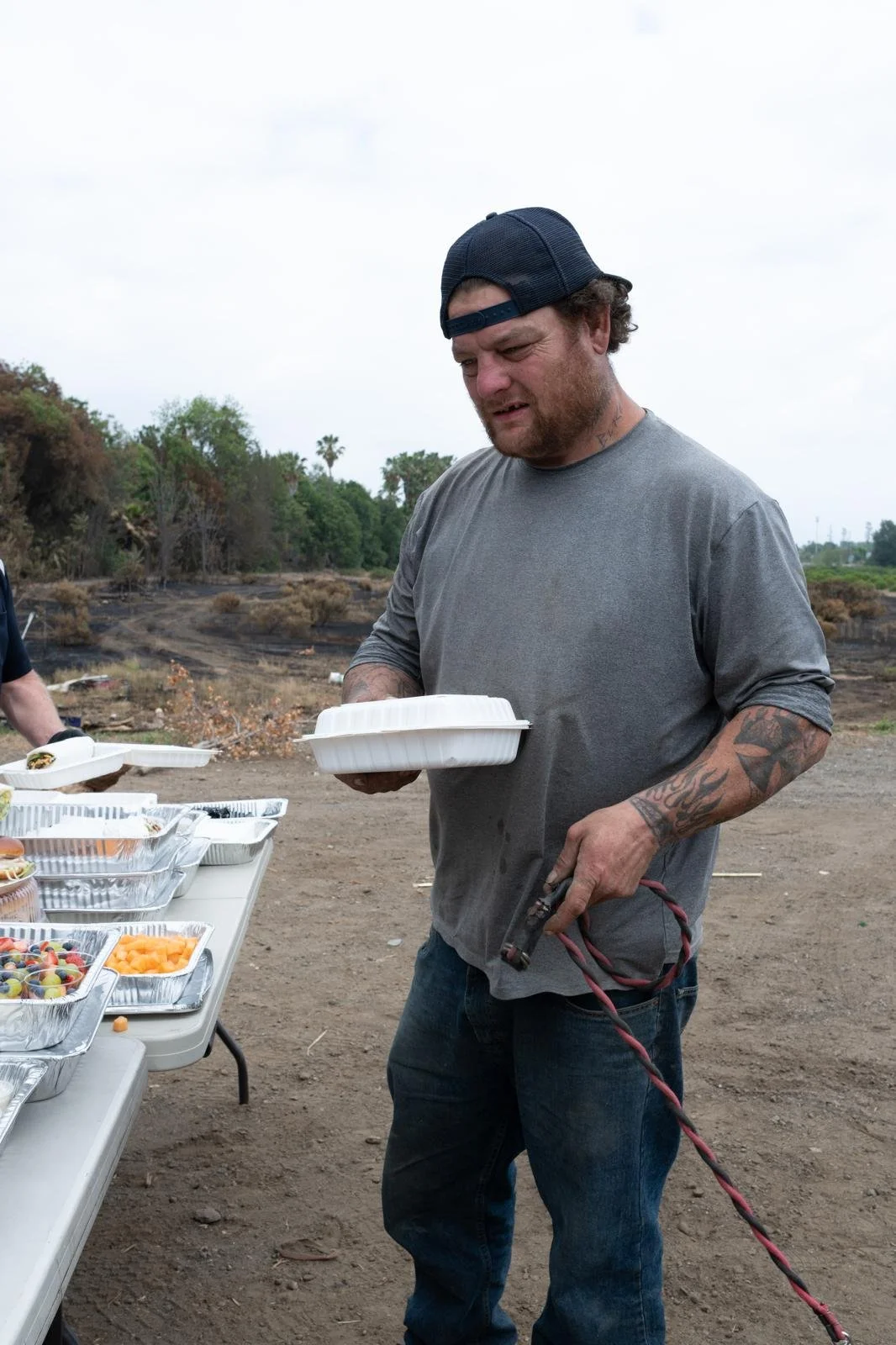 A man with tattoos on his arm, wearing a gray t-shirt and a backward baseball cap, holding a food container and a lighter, standing outdoors near a table with food containers.