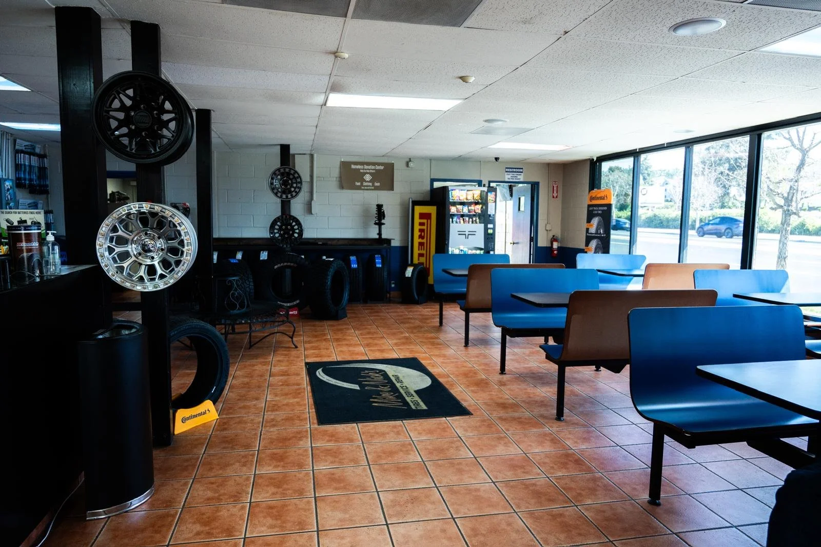 Interior of an auto service waiting area with blue and brown chairs, a vending machine, tires, car rims on display, and large windows showing trees and parked cars outside.