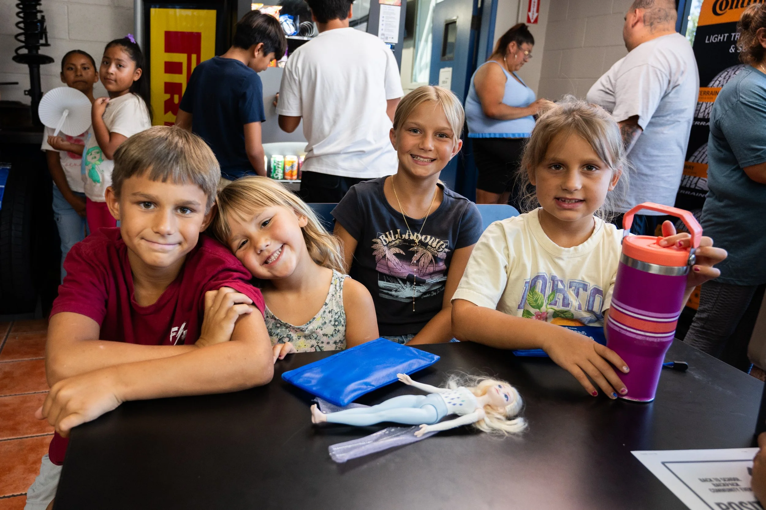 Group of children smiling and sitting at a table in a fast-food restaurant, with more people in line in the background.