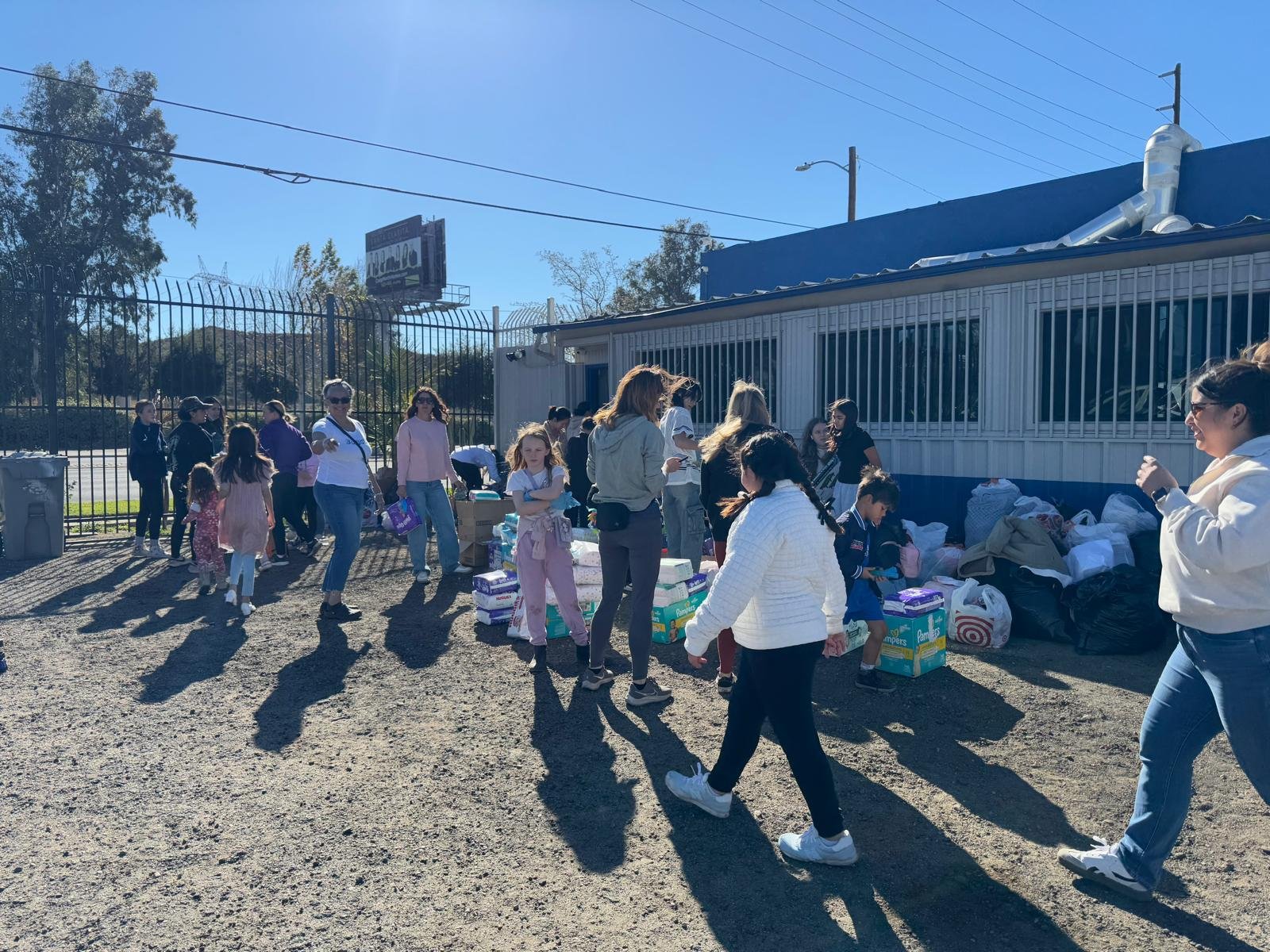 People gathering outdoors near a building, with supplies and bags on the ground, under clear blue skies.