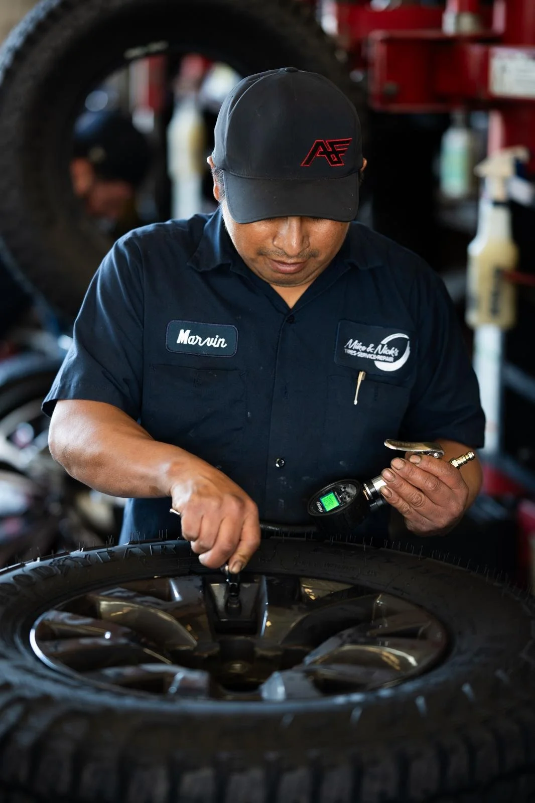 A mechanic wearing a black cap and navy blue shirt labeled 'Marvin' checks the tire pressure of a car tire in a garage.