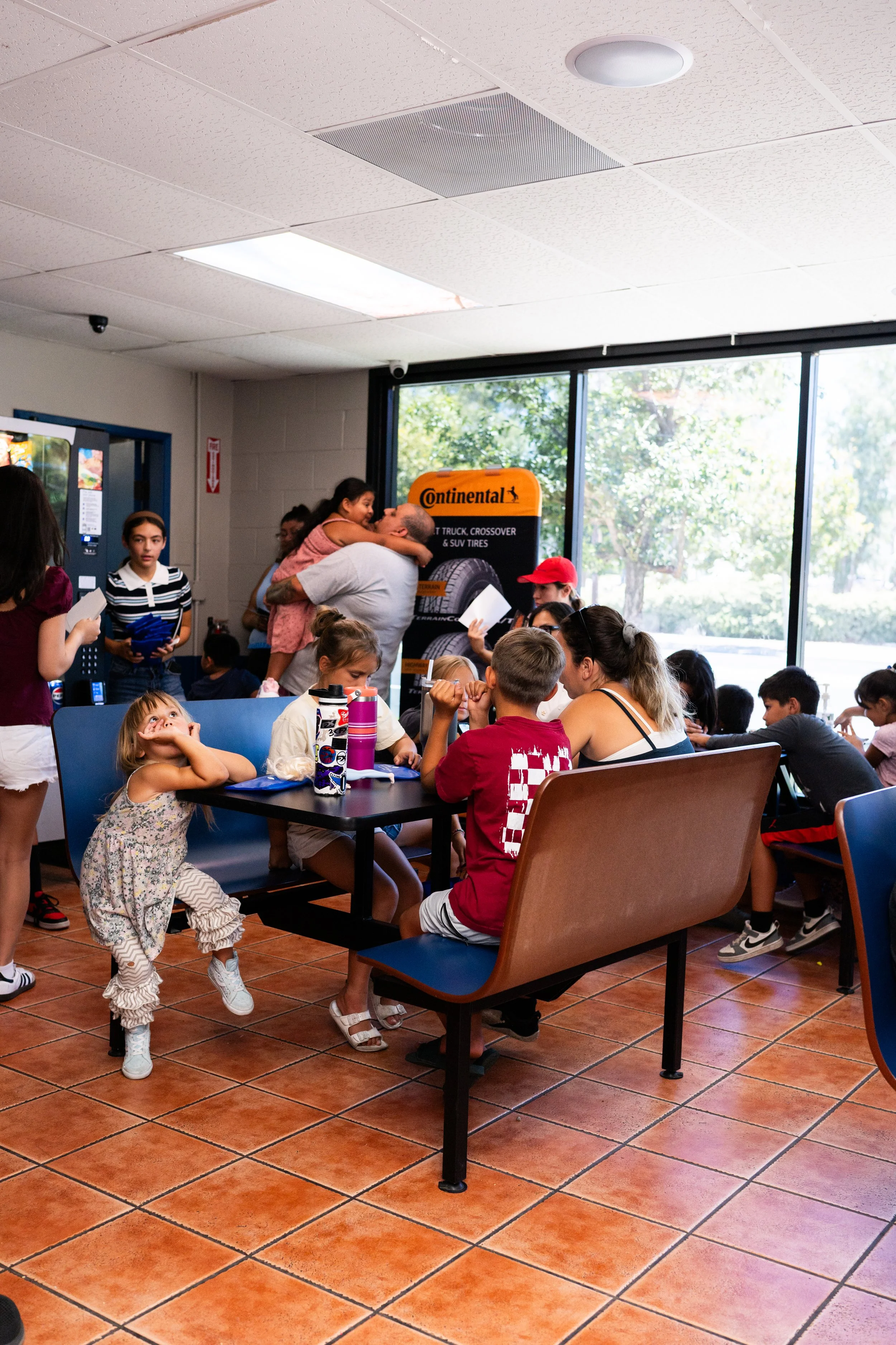 People gathered in a restaurant or cafe, sitting at tables, with a young girl standing in the foreground. Inside, a person is hugging a child, and others are engaged in conversation or activities. Large windows show trees outside, and the interior fe