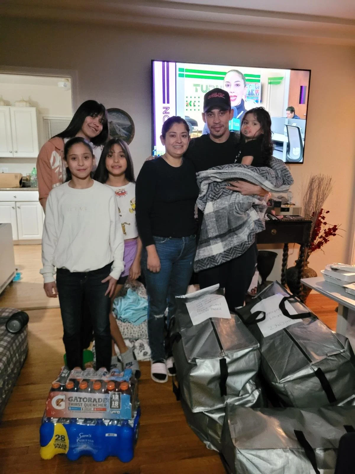 A group of six people, including children and adults, standing indoors with bags and supplies, possibly on a charitable or family gathering occasion.