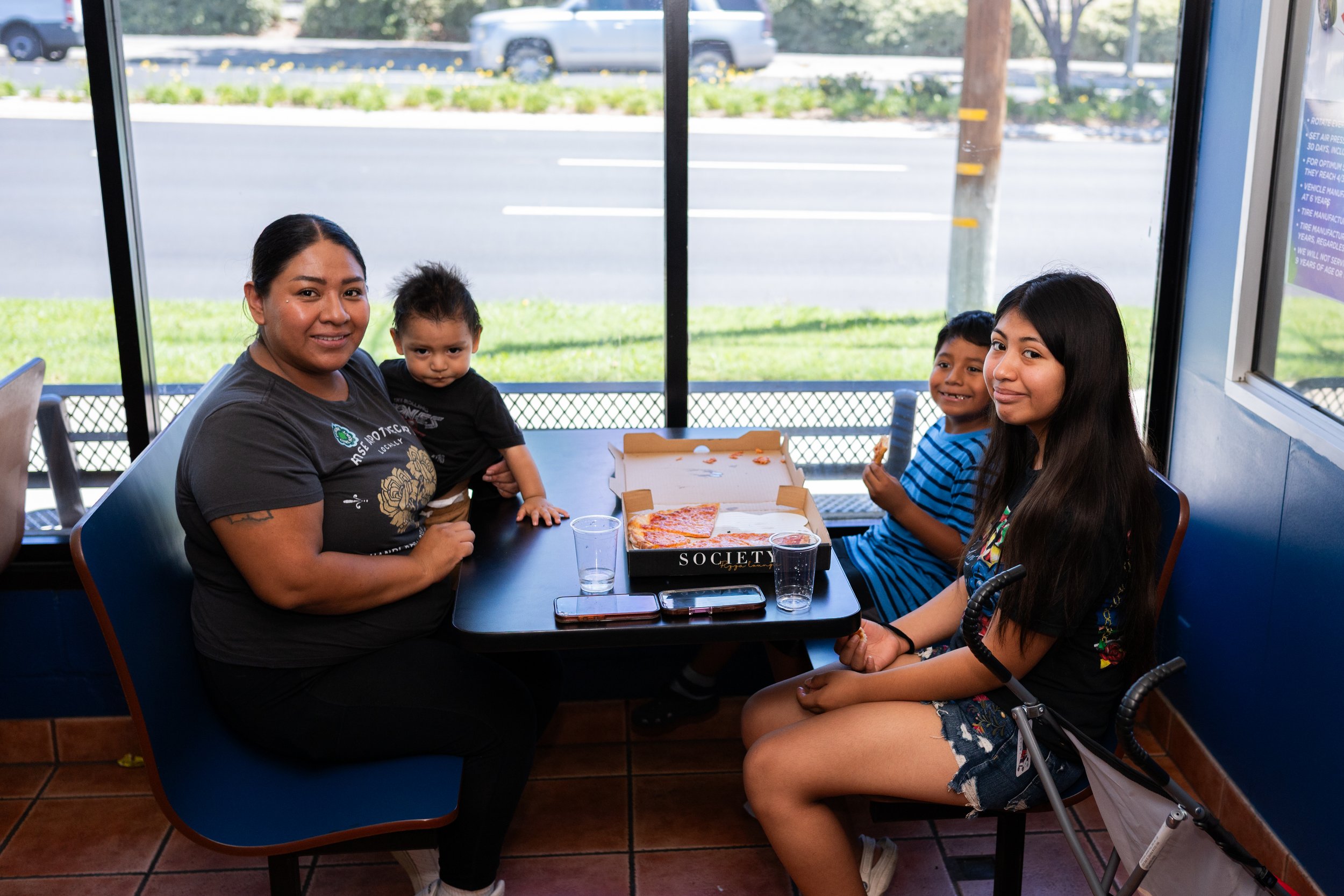 Four people sitting at a table inside a restaurant or cafe, with pizza in a box and drinks on the table, windows showing a parking lot outside.