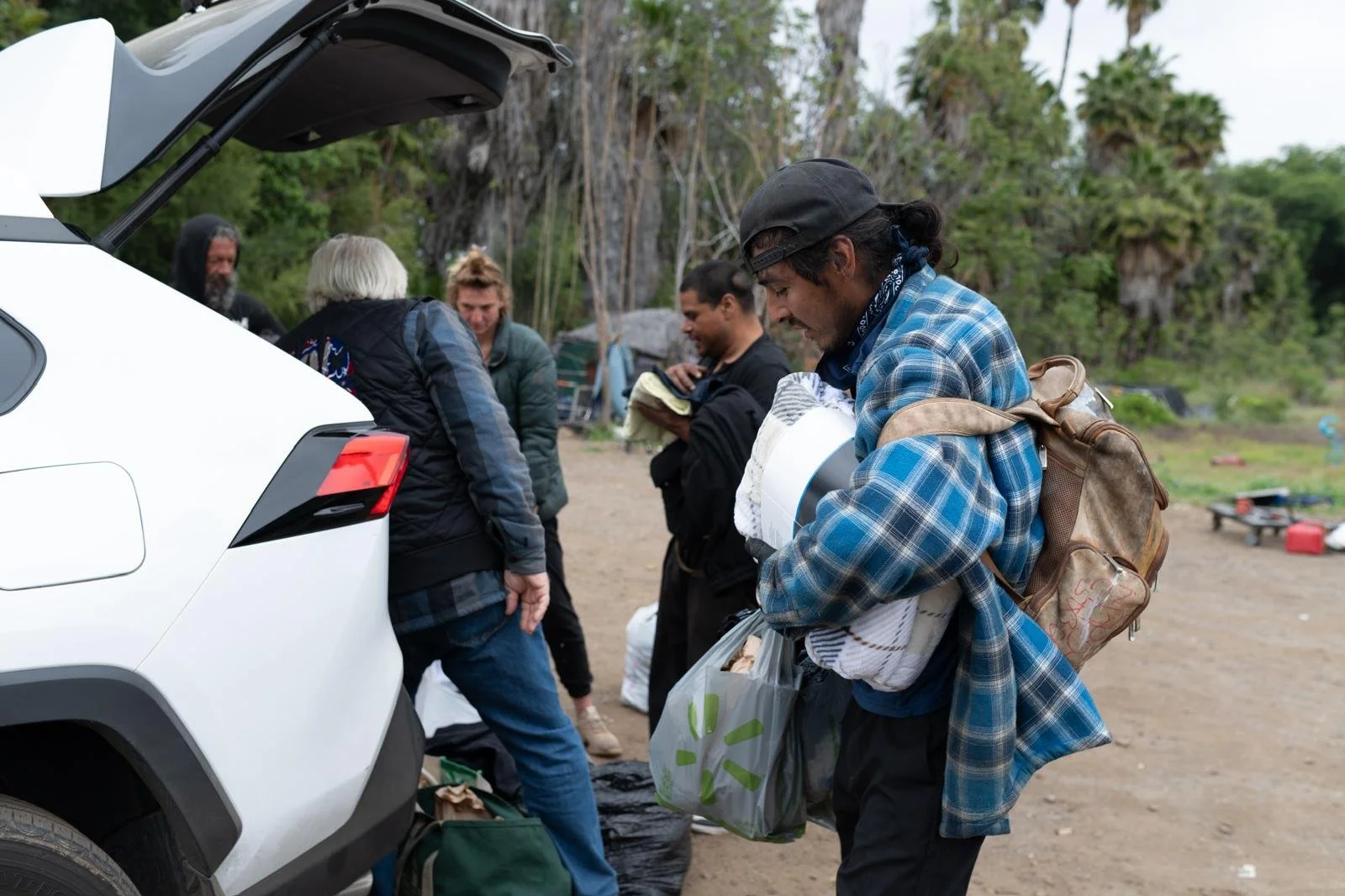 Group of people outdoors, loading items into the back of a white vehicle, with trees in the background.