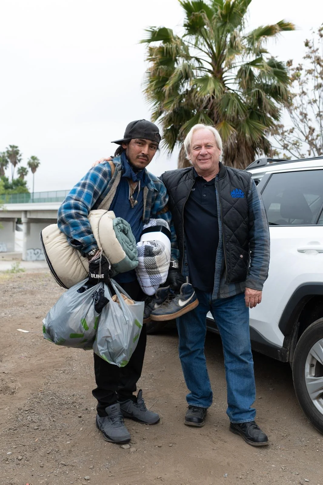 Two men standing outside near a white vehicle, with palm trees in the background. One man is holding multiple bags and shoes, while the other is resting his arm on his shoulder.