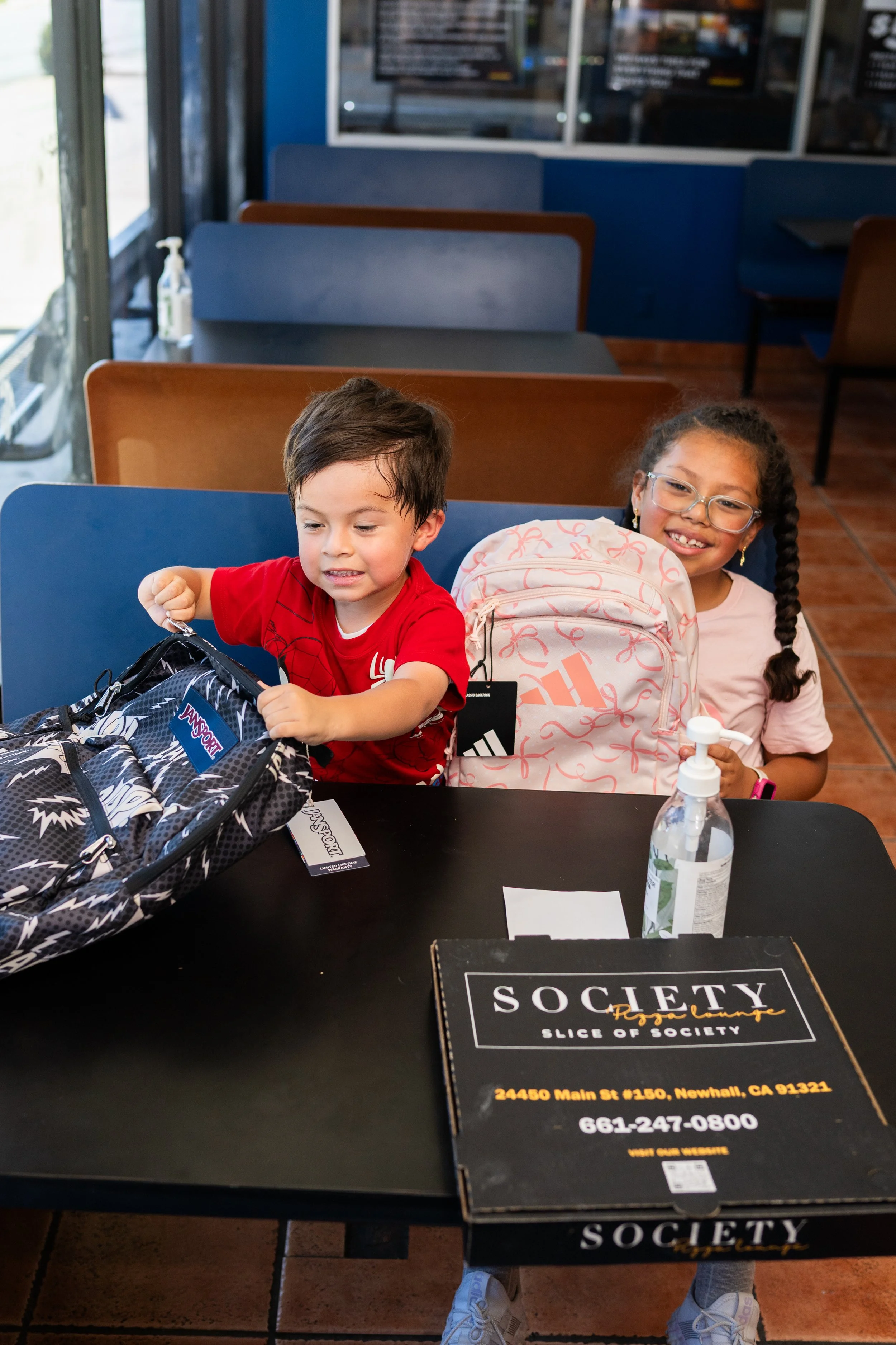 Two children sitting at a table inside a restaurant, with backpacks in front of them. The boy has short brown hair and is wearing a red shirt, while the girl has curly hair, glasses, and is smiling. There is a bottle of hand sanitizer on the table an