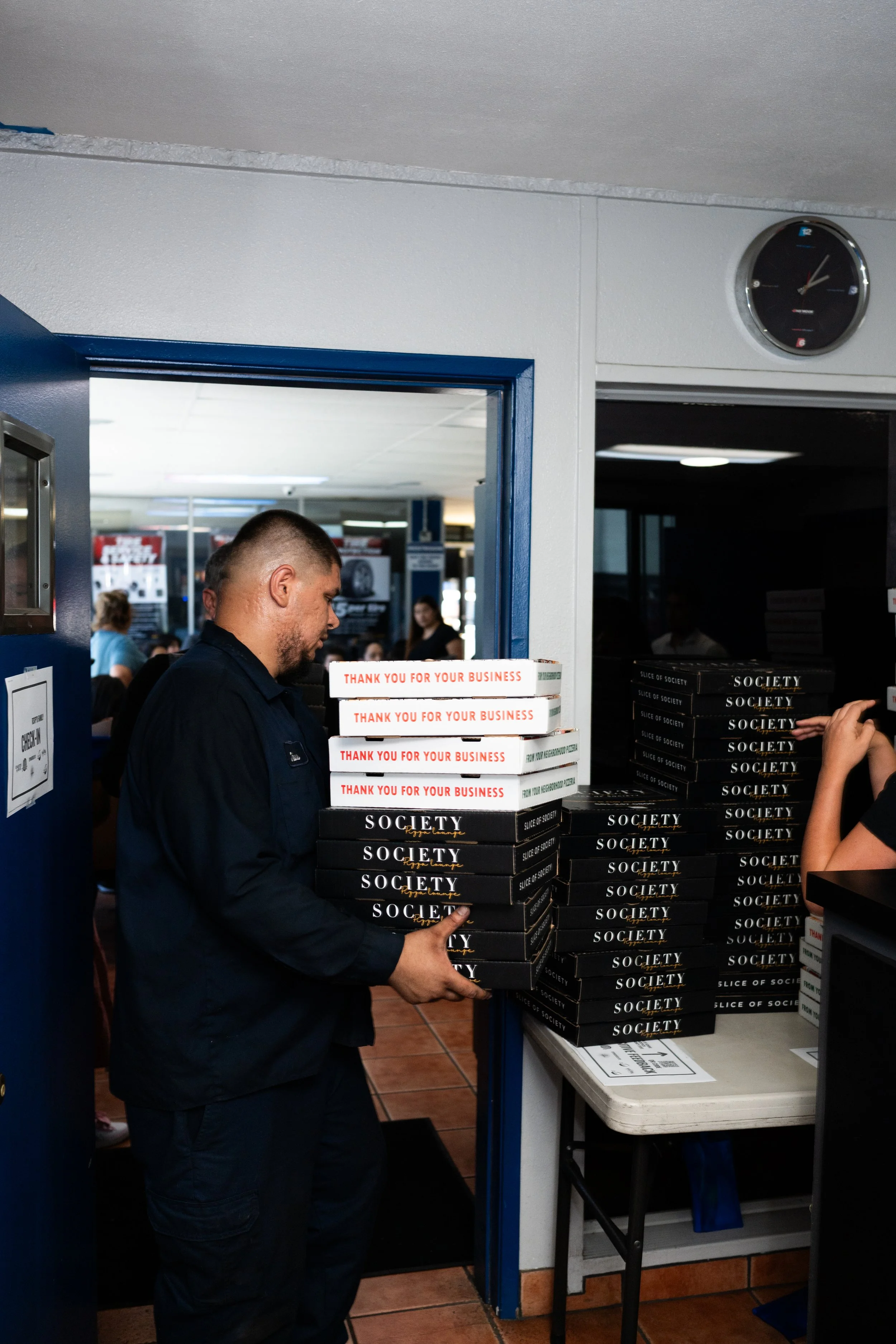A man in a black shirt is carrying several black takeaway boxes with gold and white text that says 'Society' and 'Revel Lounge'. He is inside a fast-food or restaurant setting. Behind him, there are some other people and posters on the wall. The sett