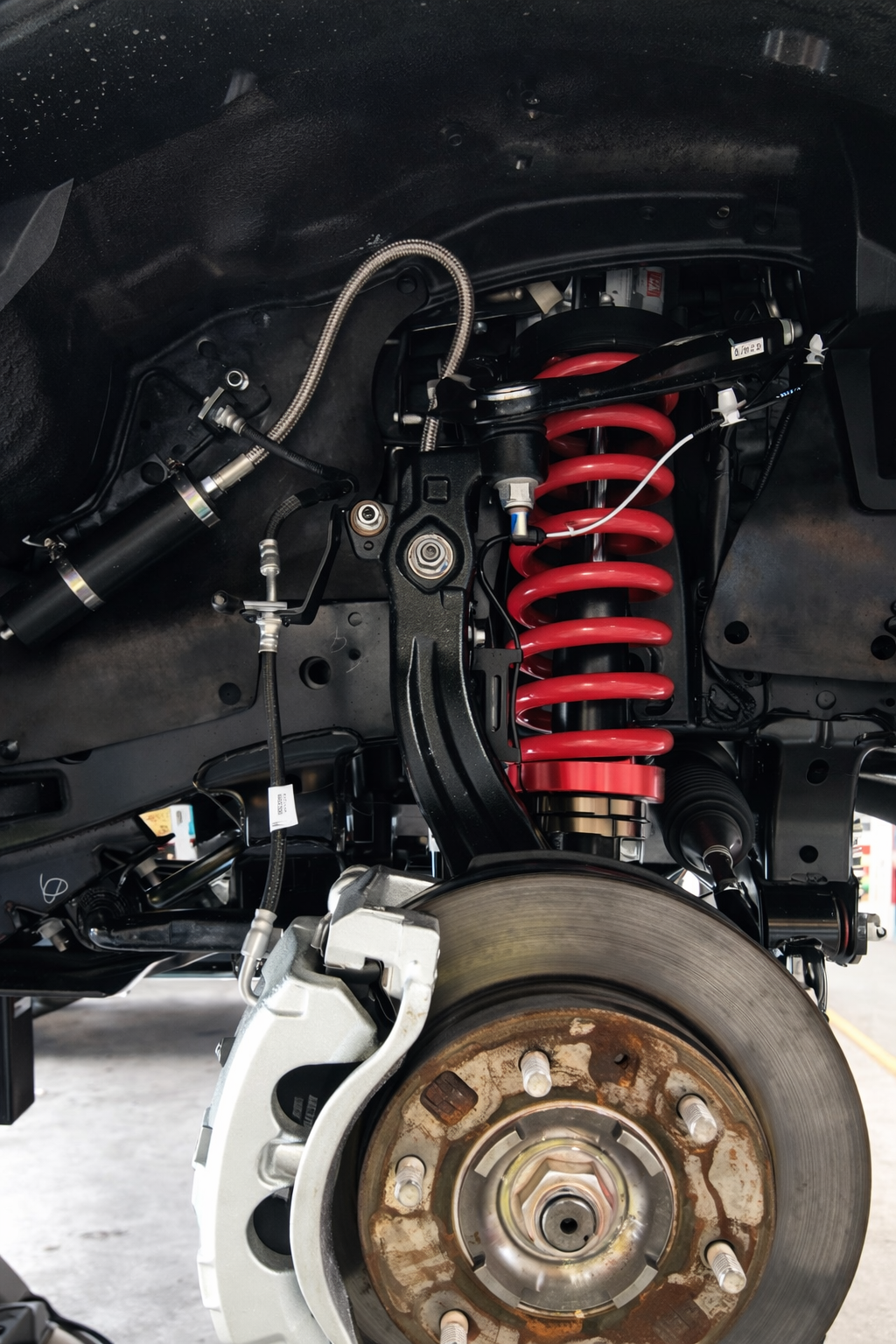 Close-up of a car's suspension system showing a red coil spring, shock absorber, brake rotor, and various mechanical components.