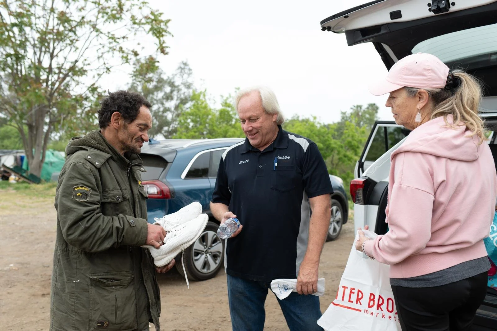 Three people standing outdoors near cars, with one man holding white shoes, and a woman holding a grocery bag, engaged in conversation.