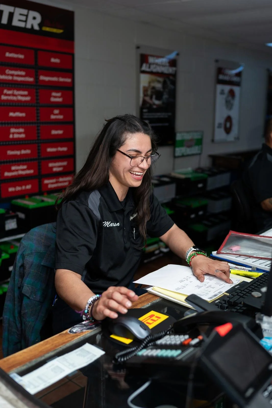 A smiling woman wearing glasses, a black polo shirt with the name 'Marsa,' sitting at a desk with files, a computer, and a phone in a service center or auto shop.
