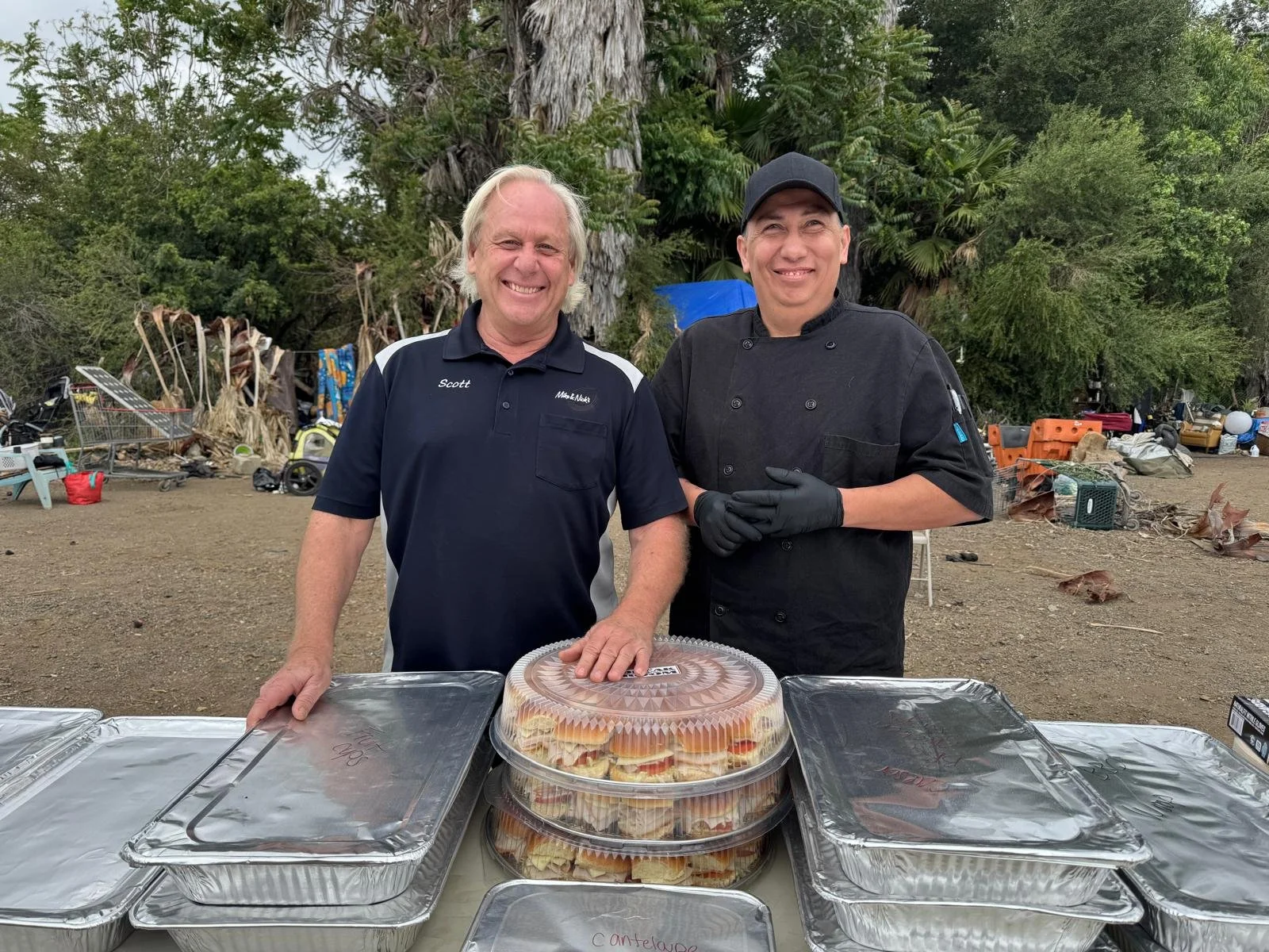 Two men standing behind a table with food containers outdoors, smiling at the camera. One man is wearing a black chef's coat and cap, the other is wearing a navy blue polo shirt.