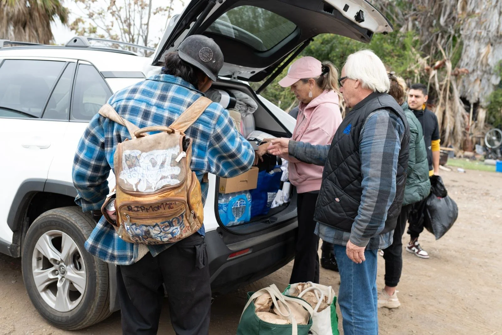 Group of people loading supplies into a vehicle on a dirt area, with trees and a wooded background.