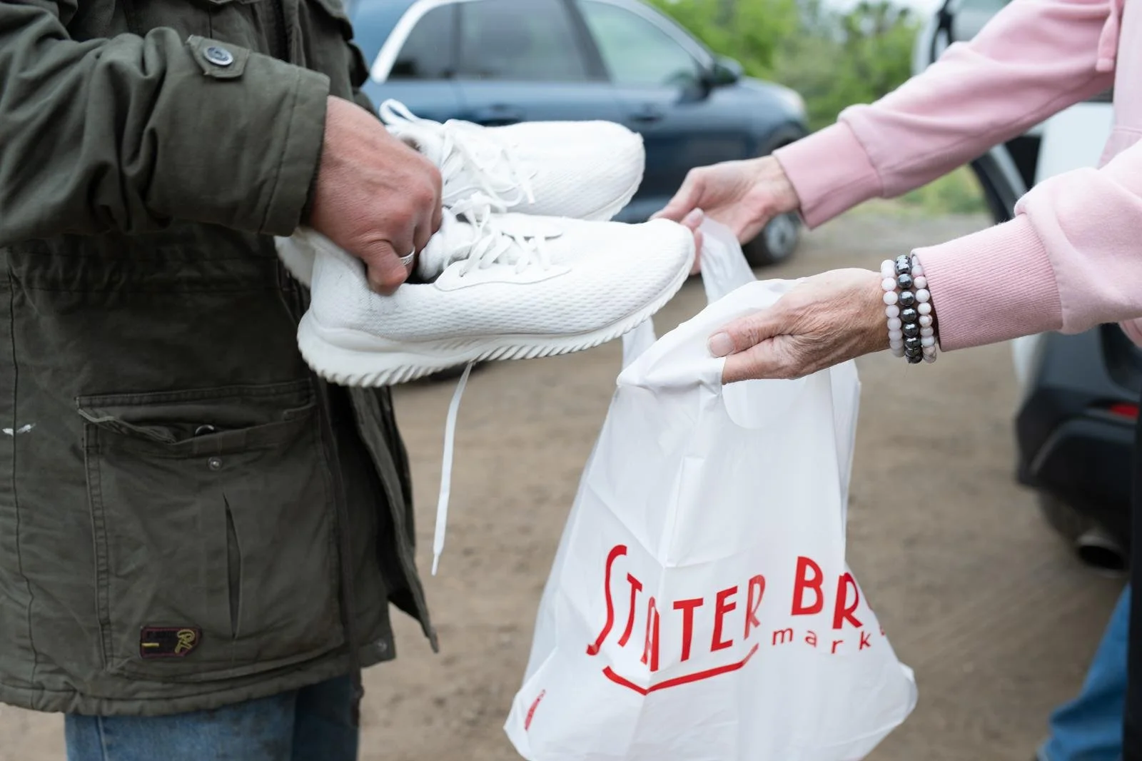 Two people exchanging a pair of white sneakers outside, near parked cars, with one person holding a white shopping bag with red text.