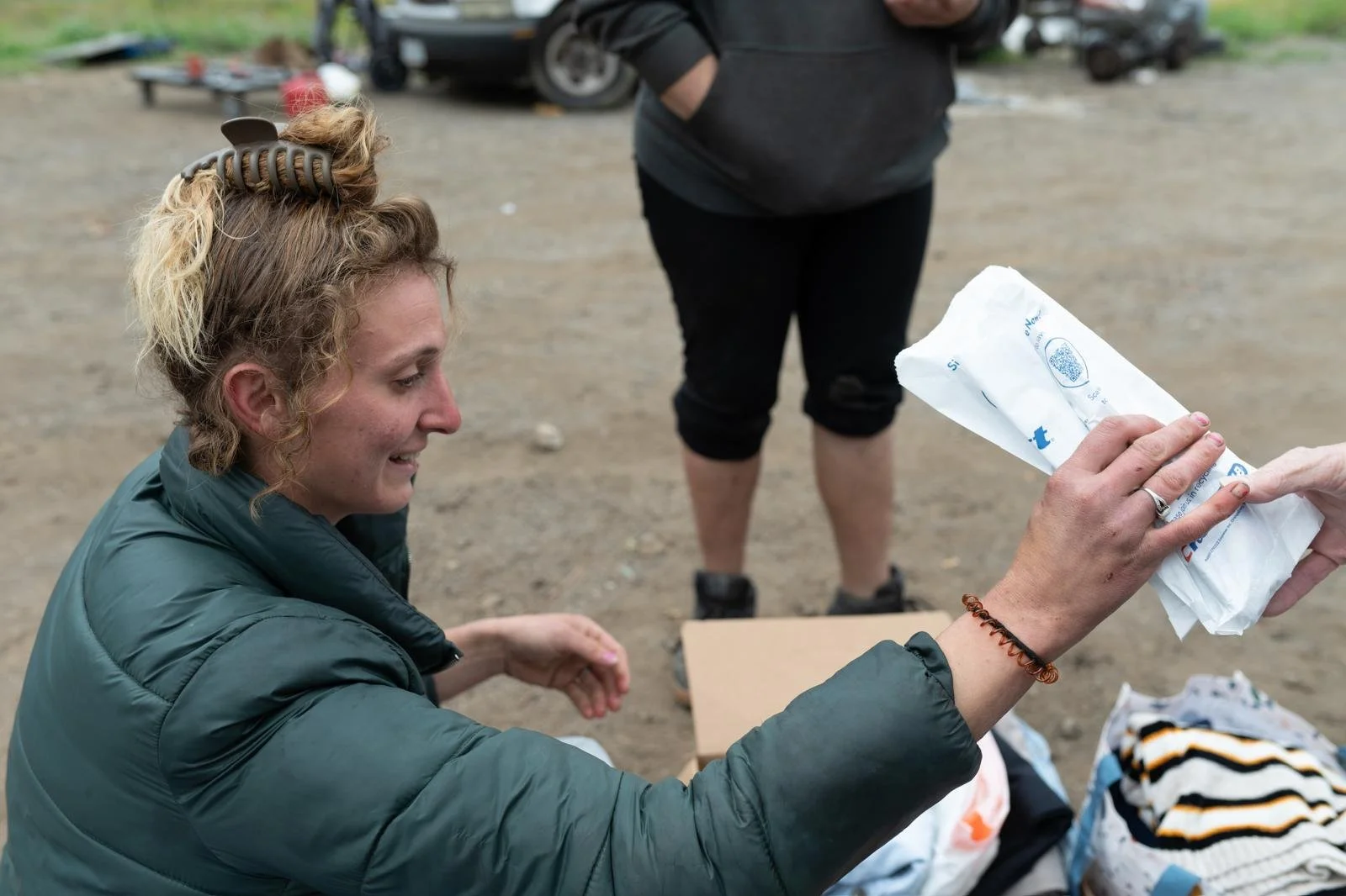 A woman with curly hair, sitting outdoors, hands outstretched, and smiling while holding a paper bag handed to her by another person. A man wearing black shorts and a hoodie stands behind her near a parked vehicle, with camping or outdoor gear nearby