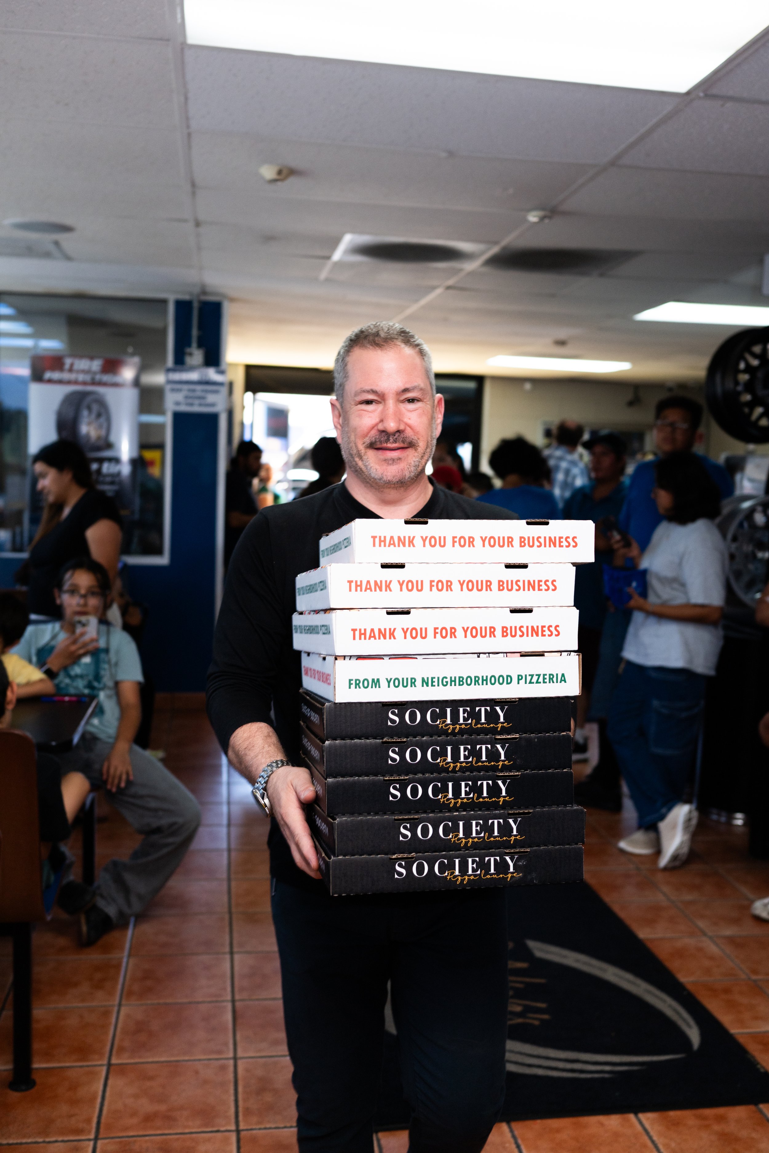 A man holding several pizza boxes with a background of people inside a restaurant or pizzeria.