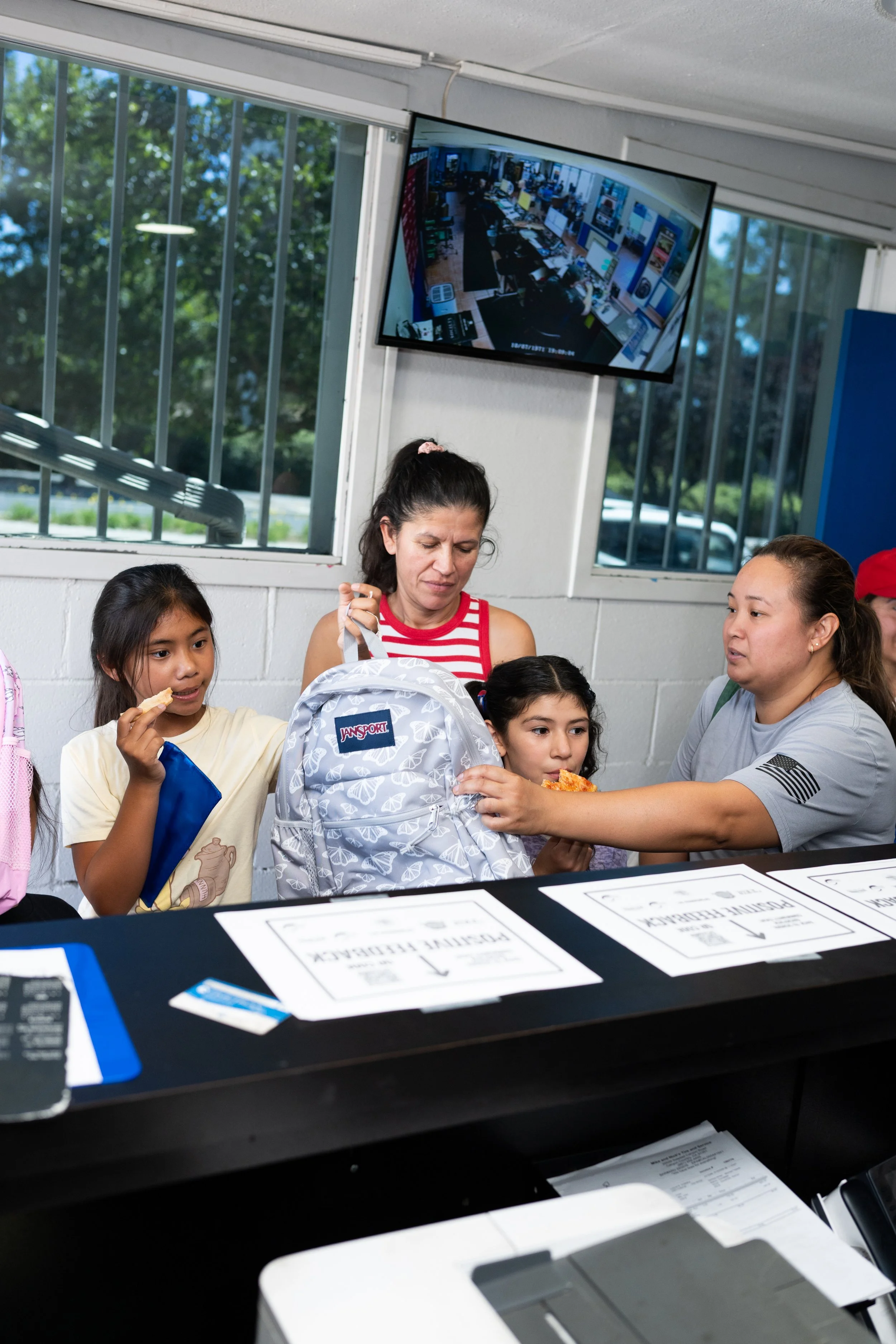A woman and children at a table, exchanging a pizza slice, with papers and a backpack nearby. A TV on the wall displays a security camera feed of the room.