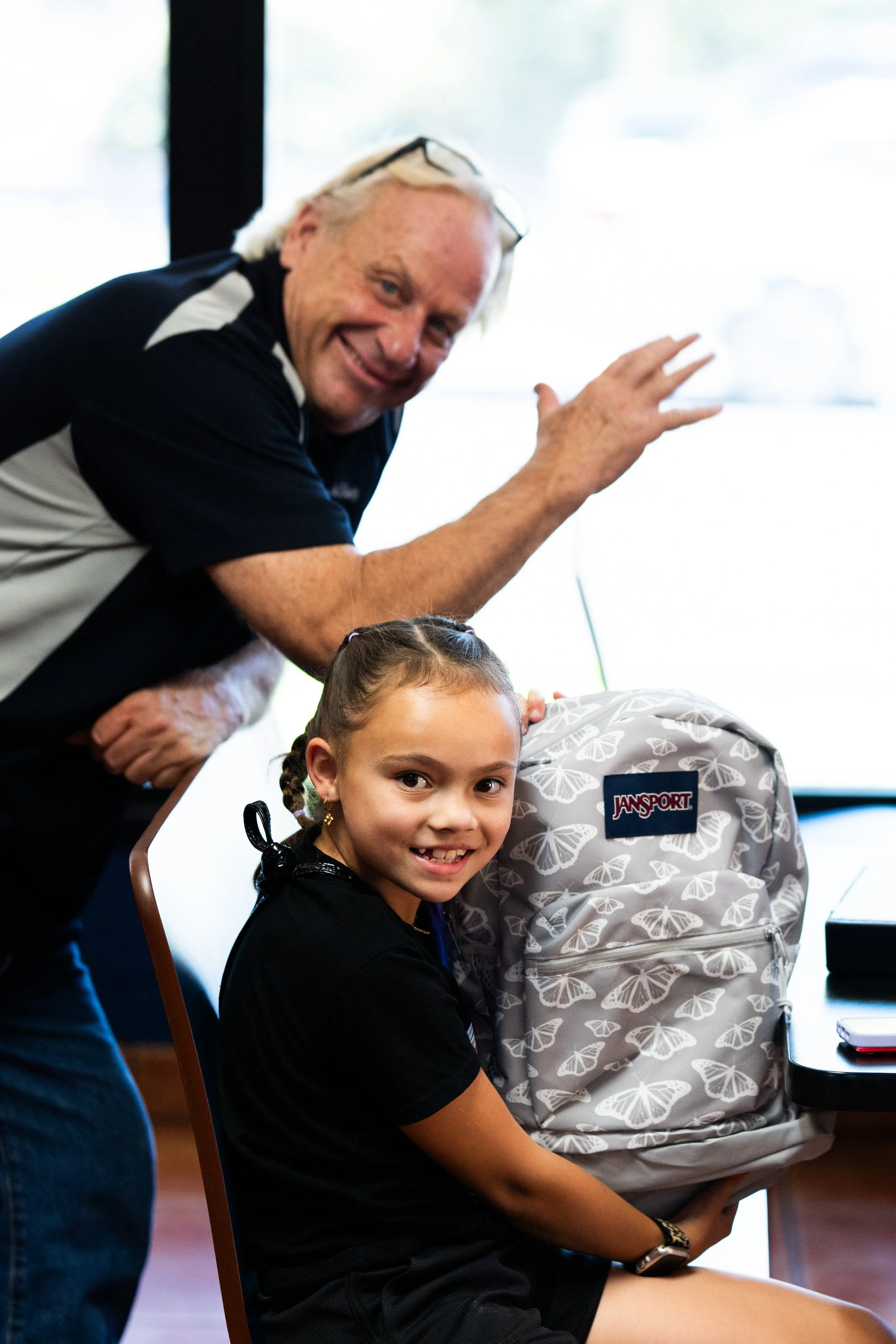 A young girl with braided hair smiling while sitting on a chair in a room, with an older man smiling and waving behind her. The girl is wearing a black shirt, and there is a gray backpack with butterfly patterns beside her. The room has a large windo