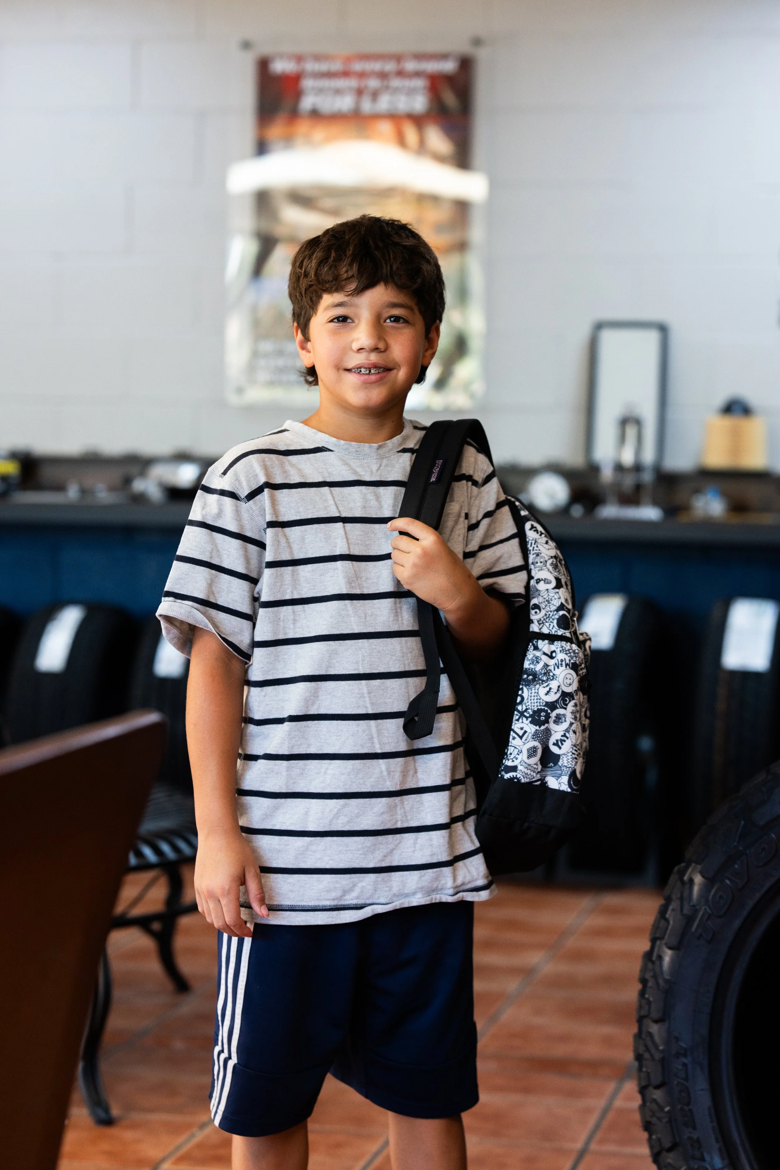 A young boy with dark hair smiling at the camera, wearing a gray and black striped t-shirt and black shorts with white stripes, standing indoors beside a large tire, holding a black backpack over one shoulder.