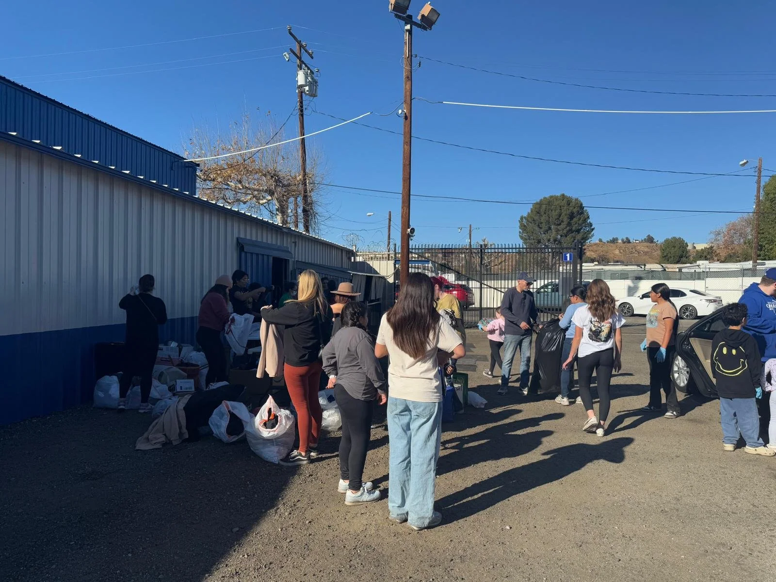 People gather outside a building, some with bags, in a line or waiting near a truck, with bags scattered on the ground, in a parking lot under a clear blue sky.