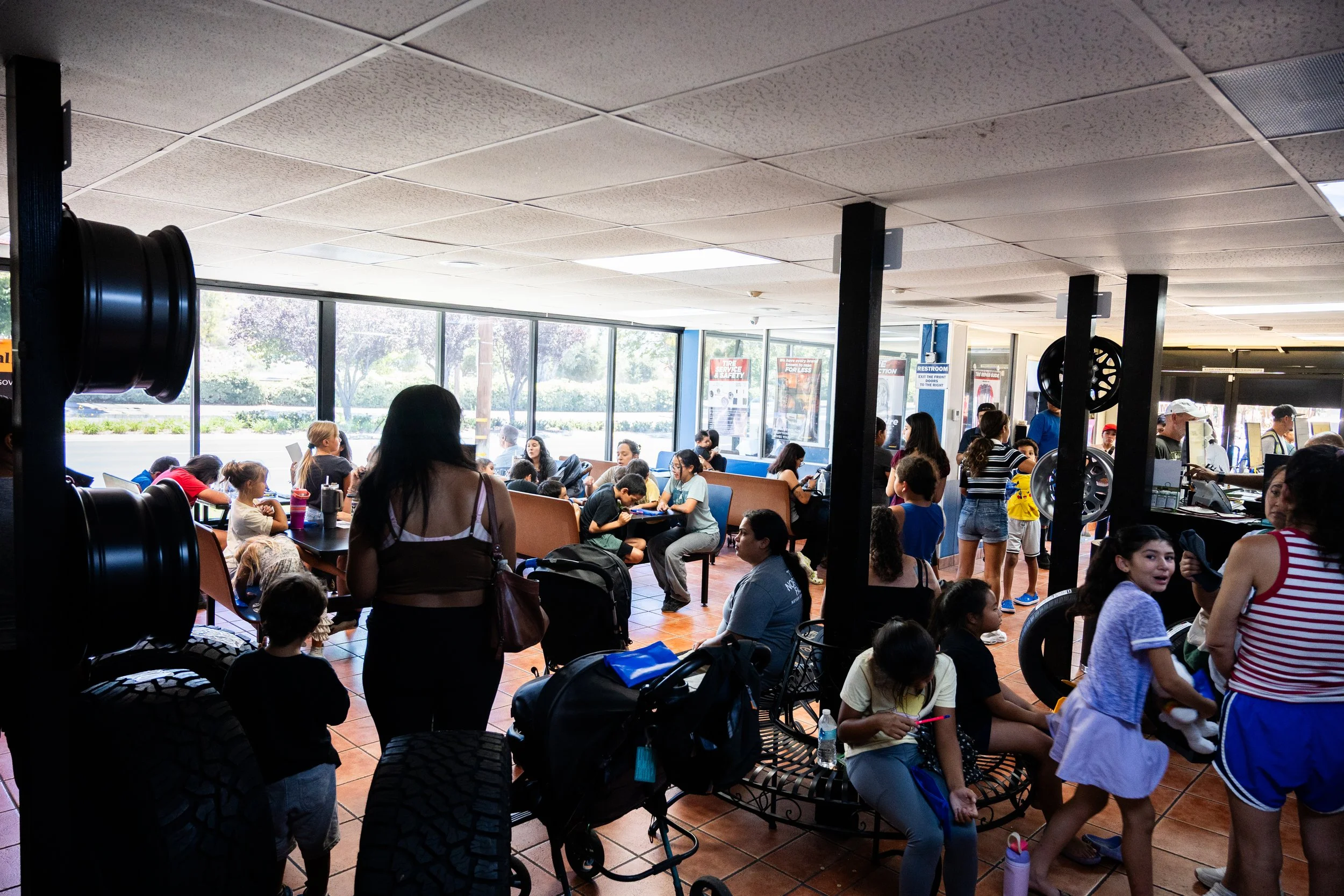 Crowded indoor waiting area with people seated and standing, large windows showing outdoor greenery, and signs on the wall, likely at a transportation hub.