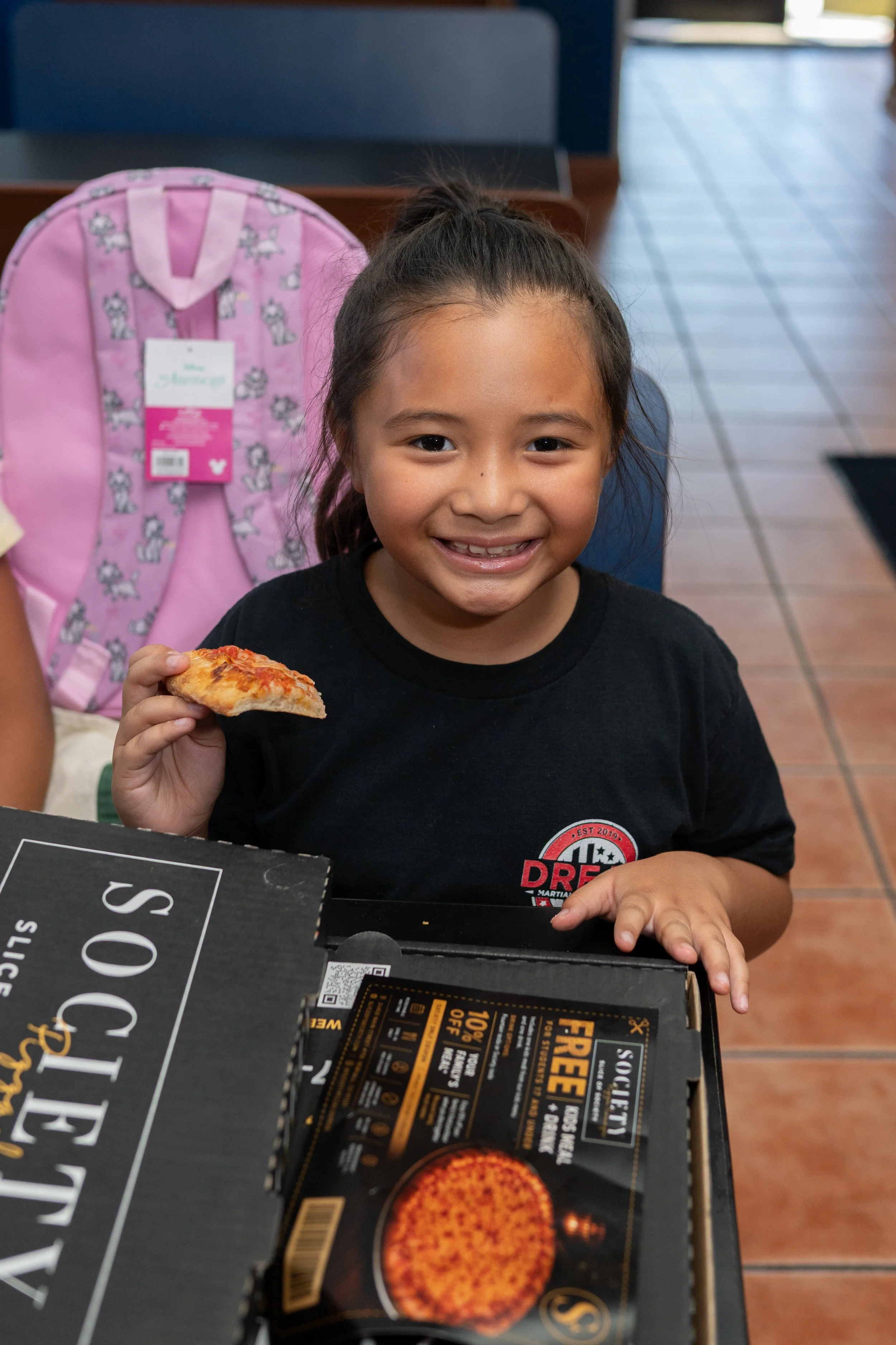 A young girl with brown hair tied in a ponytail, smiling, sitting at a table in a restaurant. She is holding a slice of pizza in her right hand, wearing a black shirt with a red and white logo. In front of her is a black box with promotional text and