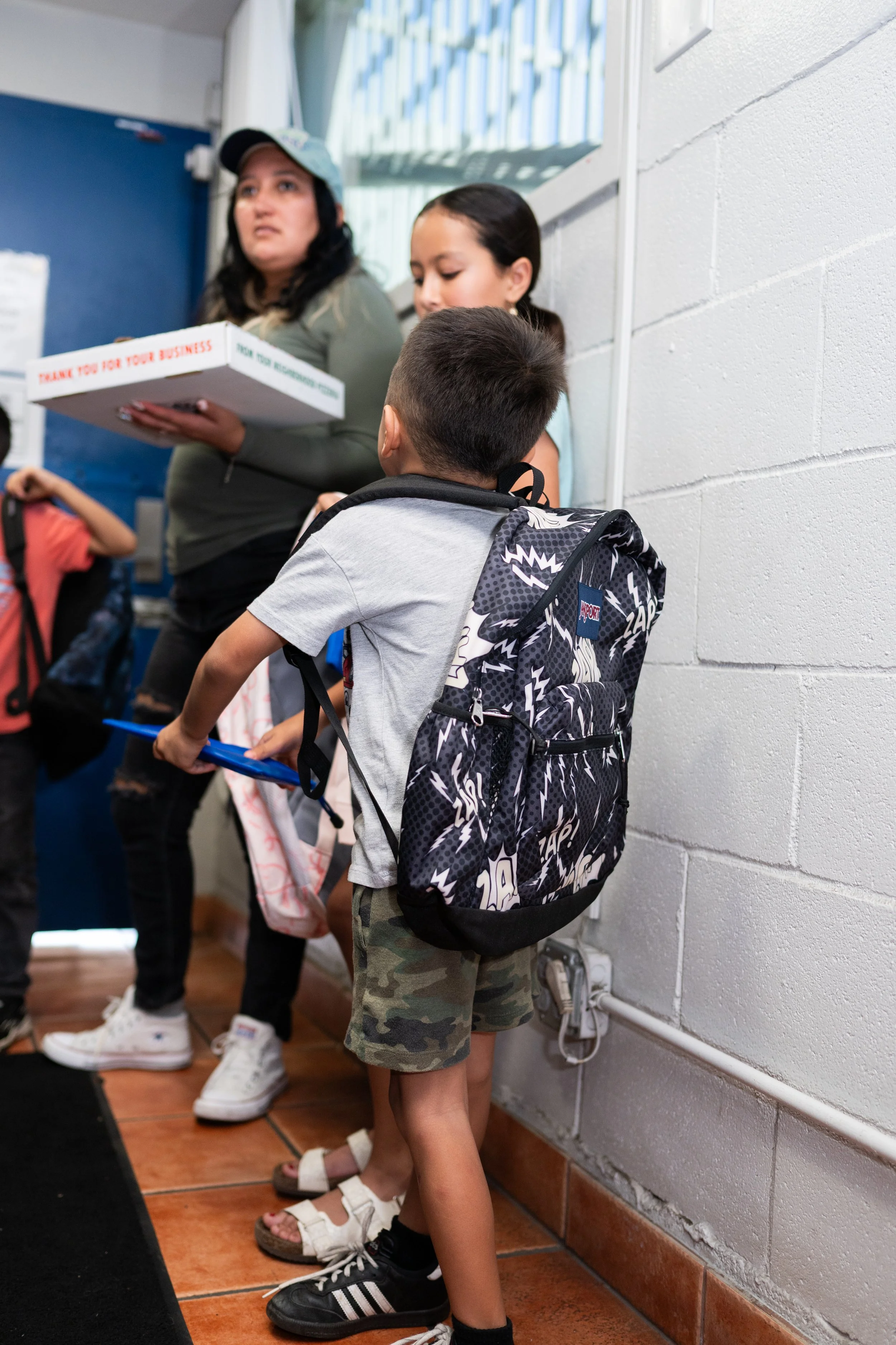 A young boy with a black and white patterned backpack, camo shorts, black sneakers, and a gray shirt stands in line against a white cinder block wall. He is looking ahead as he waits, with other children and an adult in the background, inside a schoo