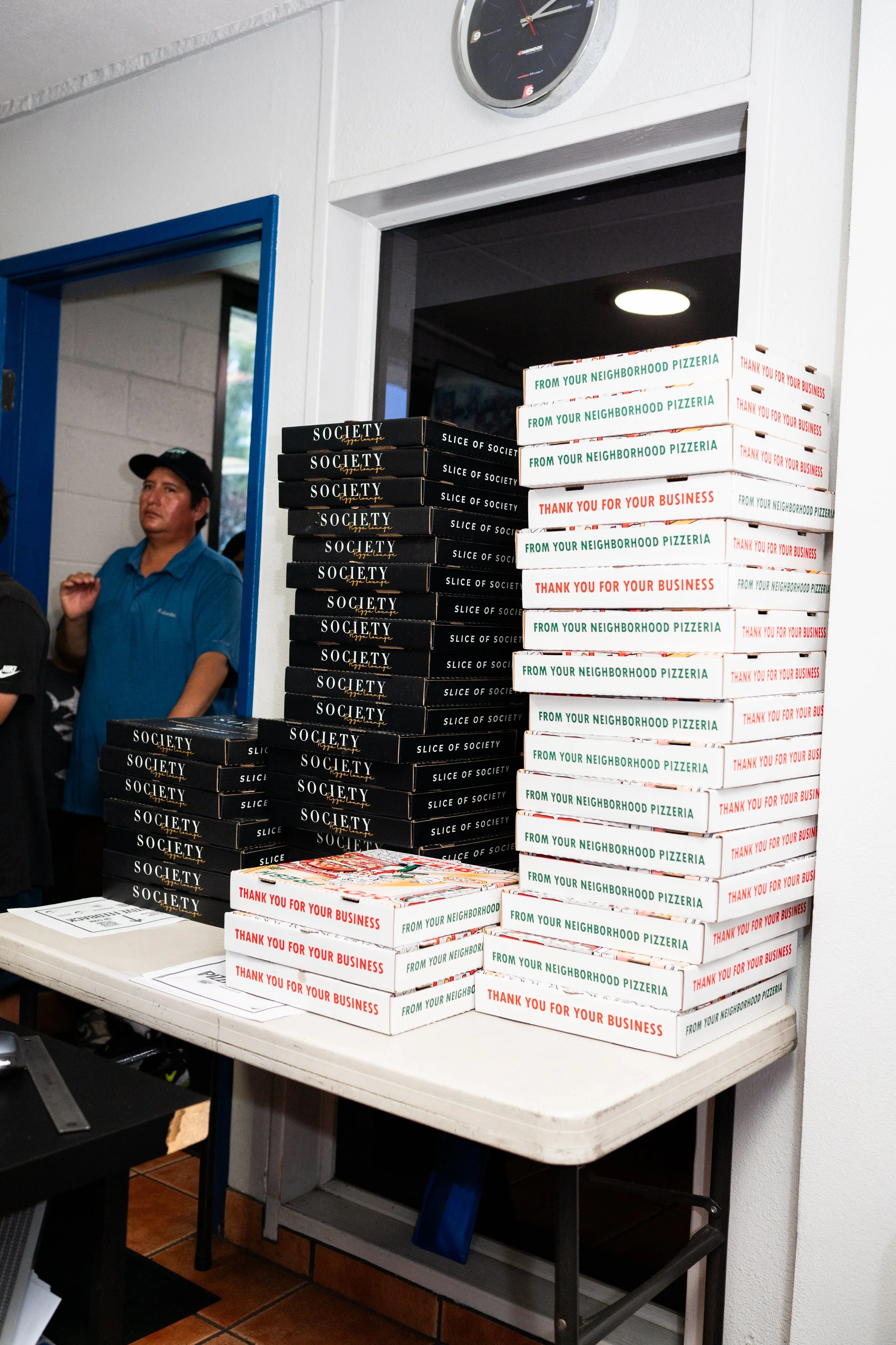 Stacks of pizza boxes on a table at a pizzeria, with a person in a blue shirt and black cap in the background.