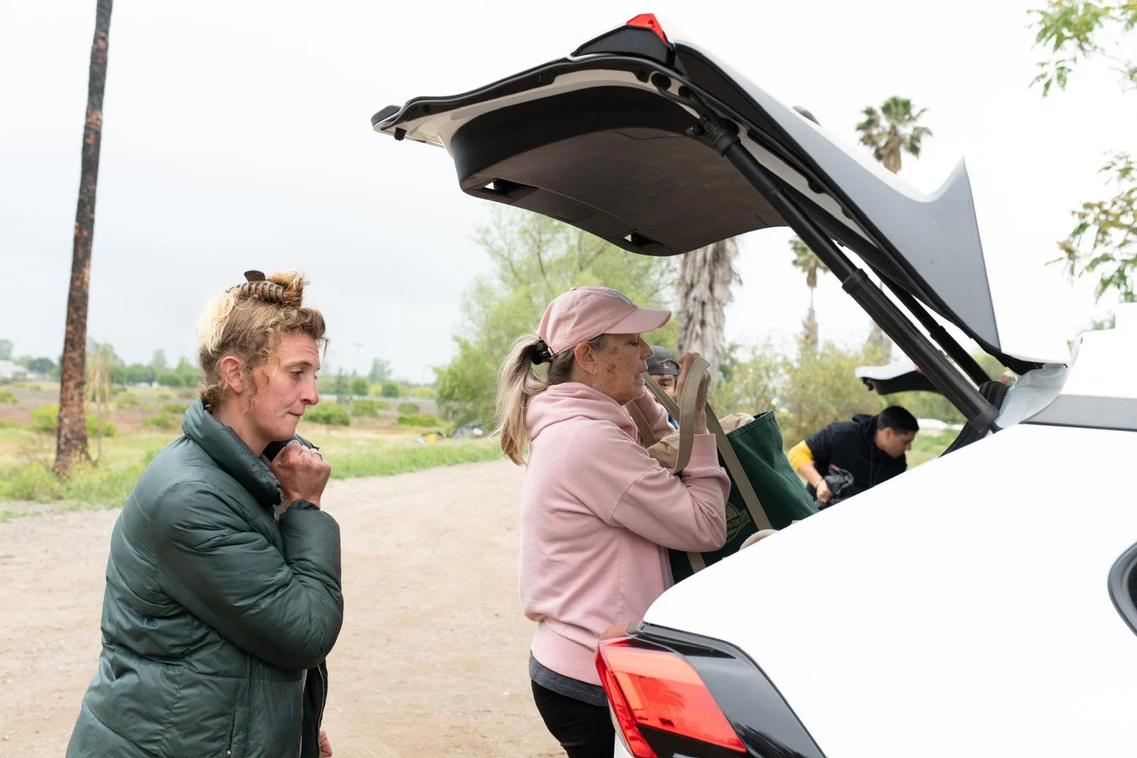 Group of people gathered around an open car trunk outdoors during the daytime, with trees and a cloudy sky in the background.