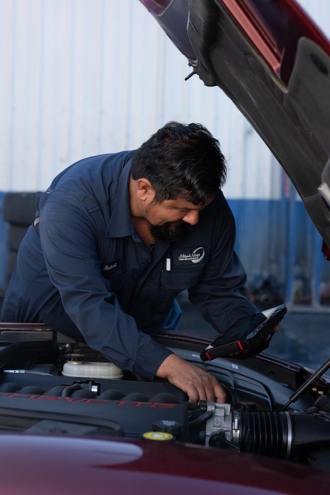A mechanic working on a red car engine with the hood open, using a diagnostic scanner in a garage.