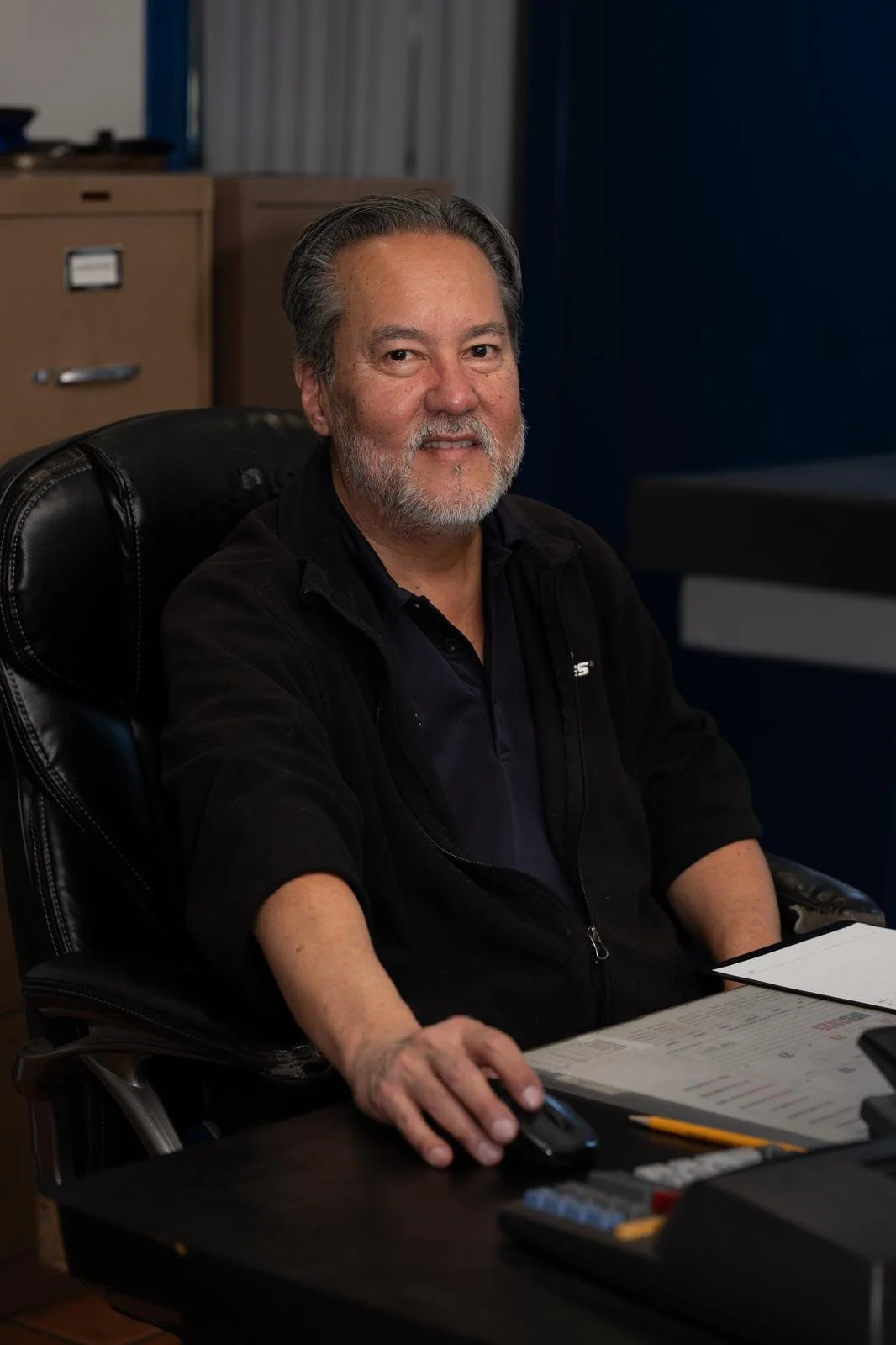 A middle-aged man with gray hair and beard, wearing a black jacket, sitting at a desk, holding a computer mouse in his right hand, smiling at the camera in an office setting.