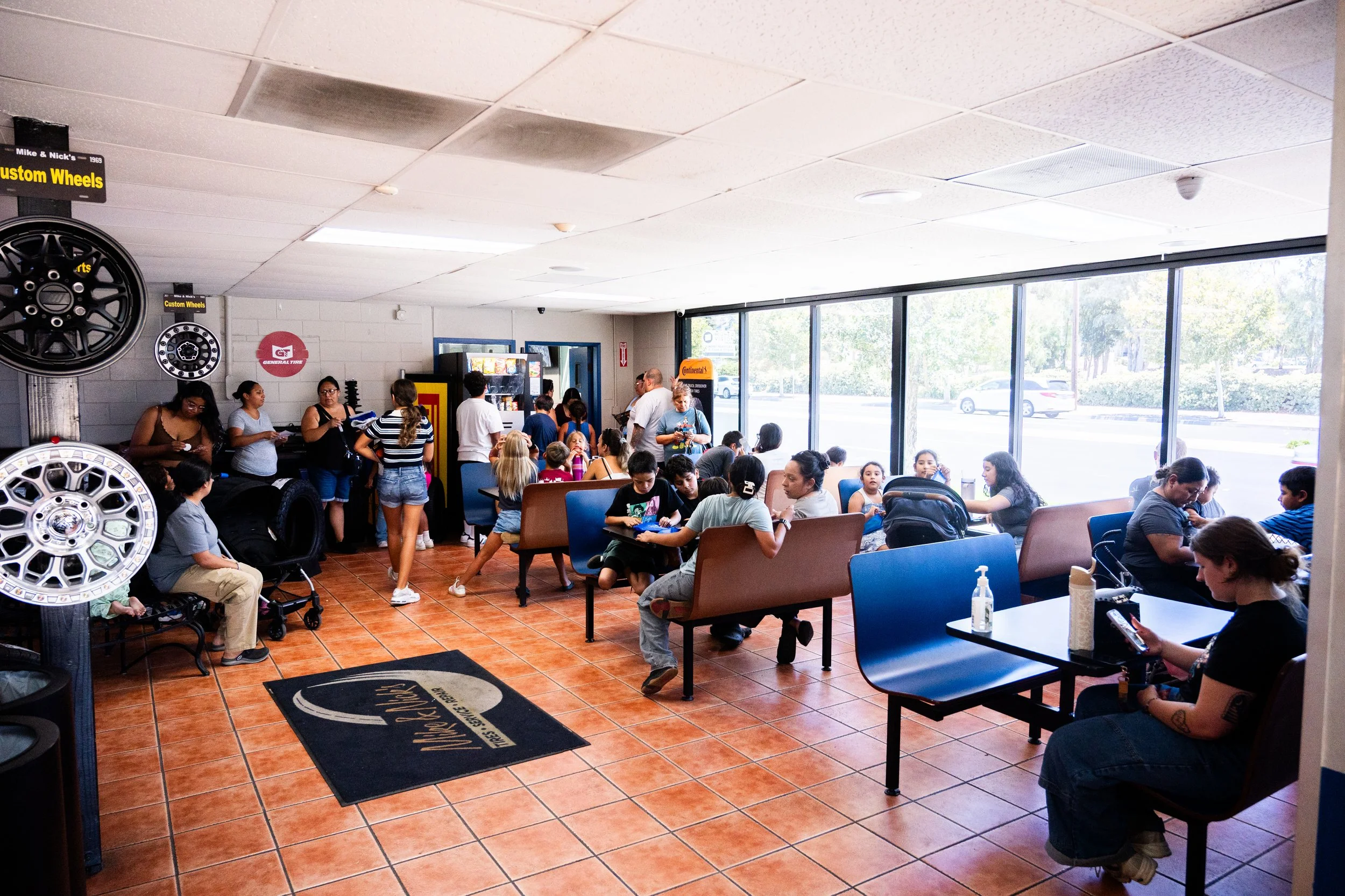 A busy auto repair shop waiting area filled with people sitting and standing, near large windows with natural light, with tires and wheels displayed on the walls.